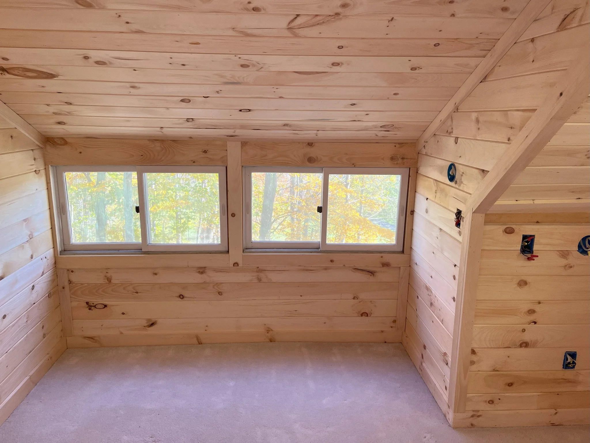 Empty room with wooden panel walls and ceiling, three large windows showing trees with autumn leaves, during construction with electrical outlets and wiring visible.