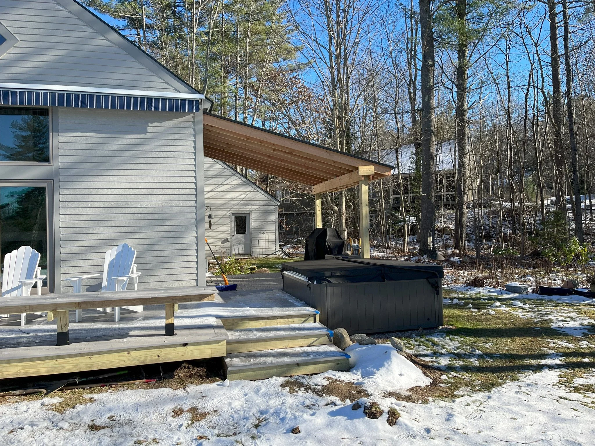 A backyard deck with two white Adirondack chairs, a hot tub, and a partially built wooden canopy in a snowy landscape with trees and a house in the background.