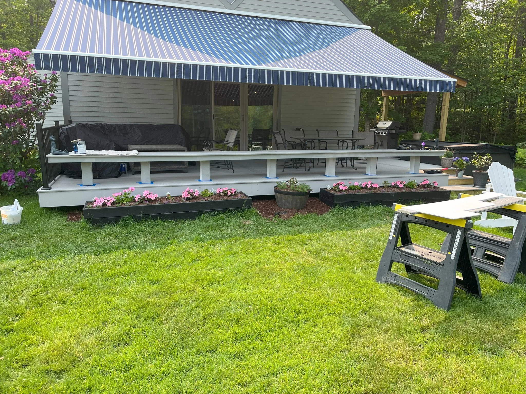 A backyard deck under construction attached to a house with a blue striped awning. There are pink flowers in black planters and a few larger pots. Construction tools and materials are present, including a saw on a stand.