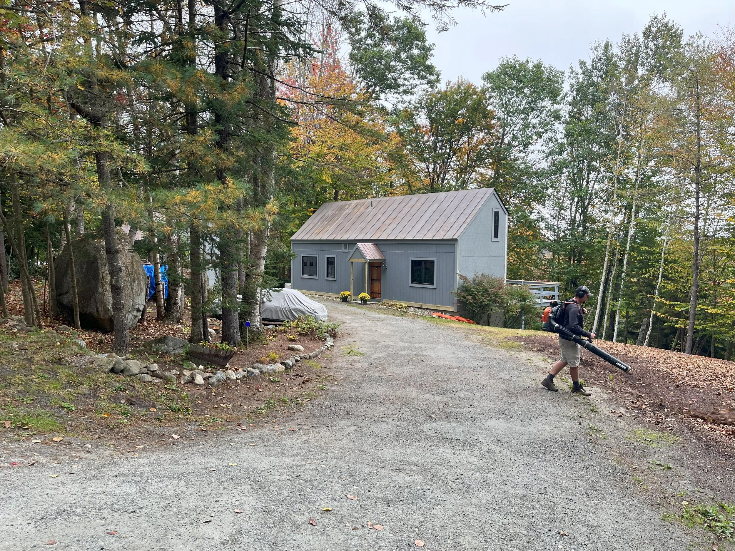 A person wearing a black hoodie, shorts, and a baseball cap using a leaf blower on a gravel driveway near a gray house with a wooden door, surrounded by trees with autumn foliage.
