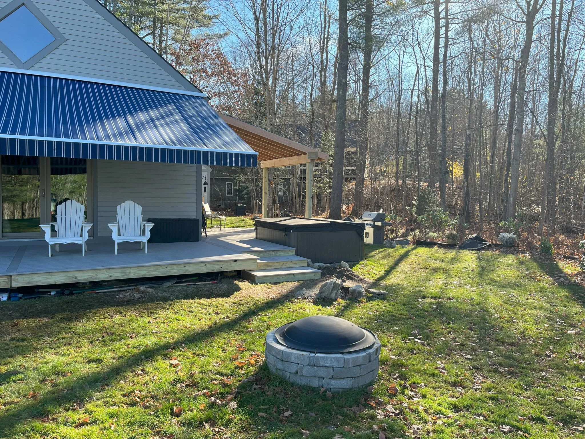 Backyard with a house and a deck, outdoor furniture, a hot tub, a grill, and a fire pit surrounded by trees and grass.