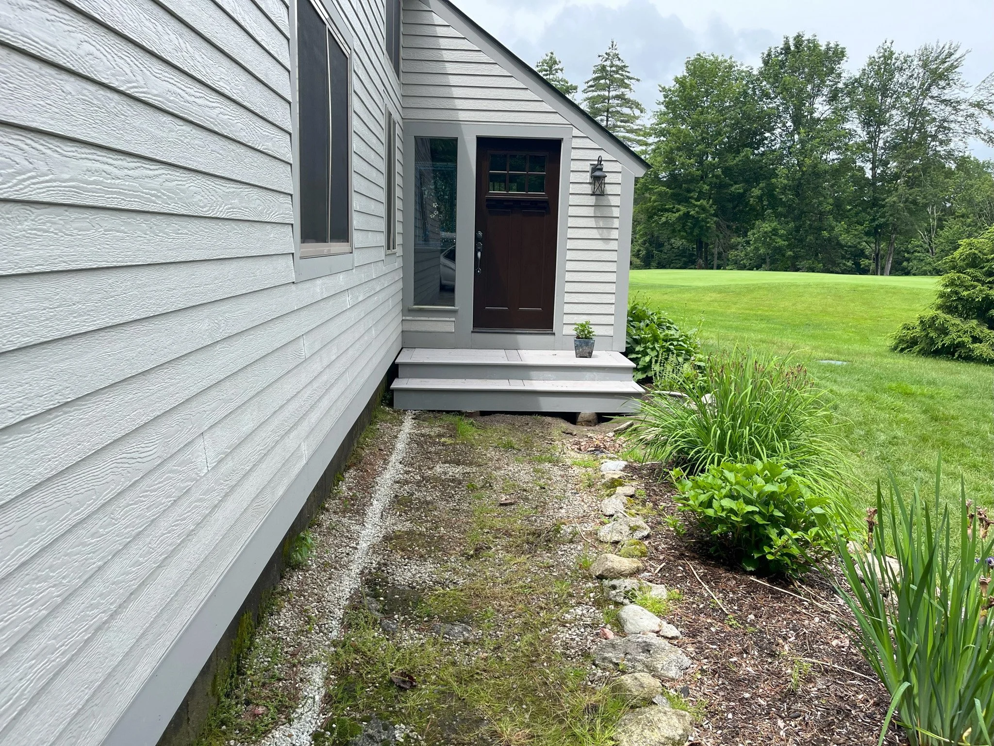 View of the porch and front door of a house with white horizontal siding, small steps leading to the door, a potted plant on the porch, and a landscaped garden with plants and rocks beside the house, with a grassy yard and trees in the background.