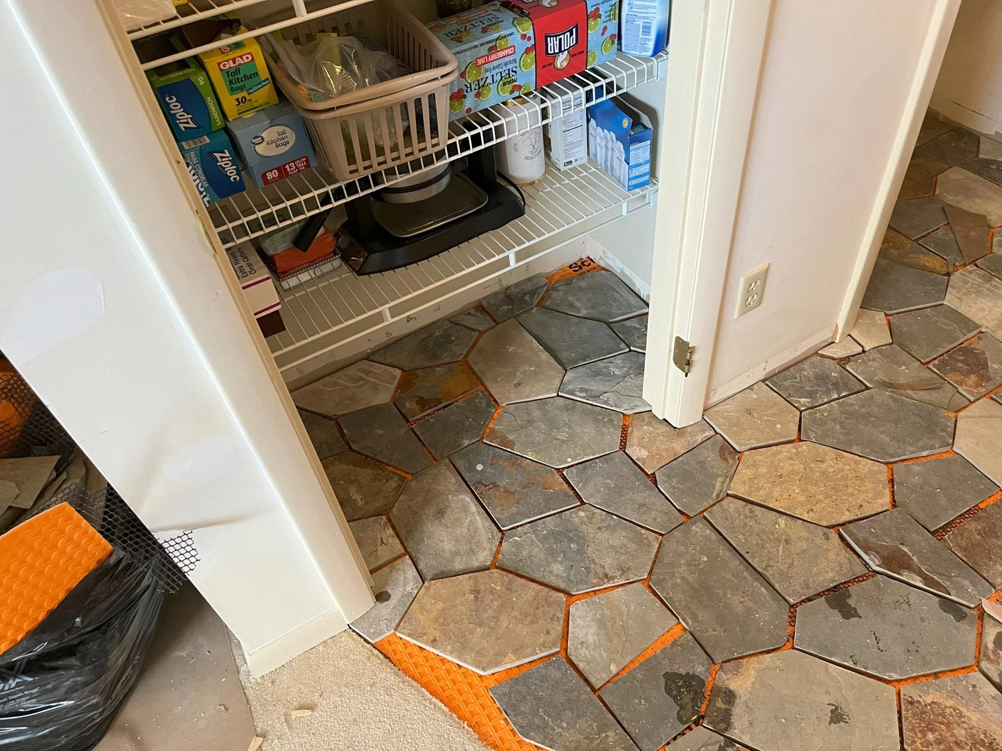 A drywall corner with a partially completed stone tile floor pattern. The pattern features large irregularly shaped stone tiles with orange grout, extending into an adjacent area with a different type of floor. The open pantry door reveals shelves wi