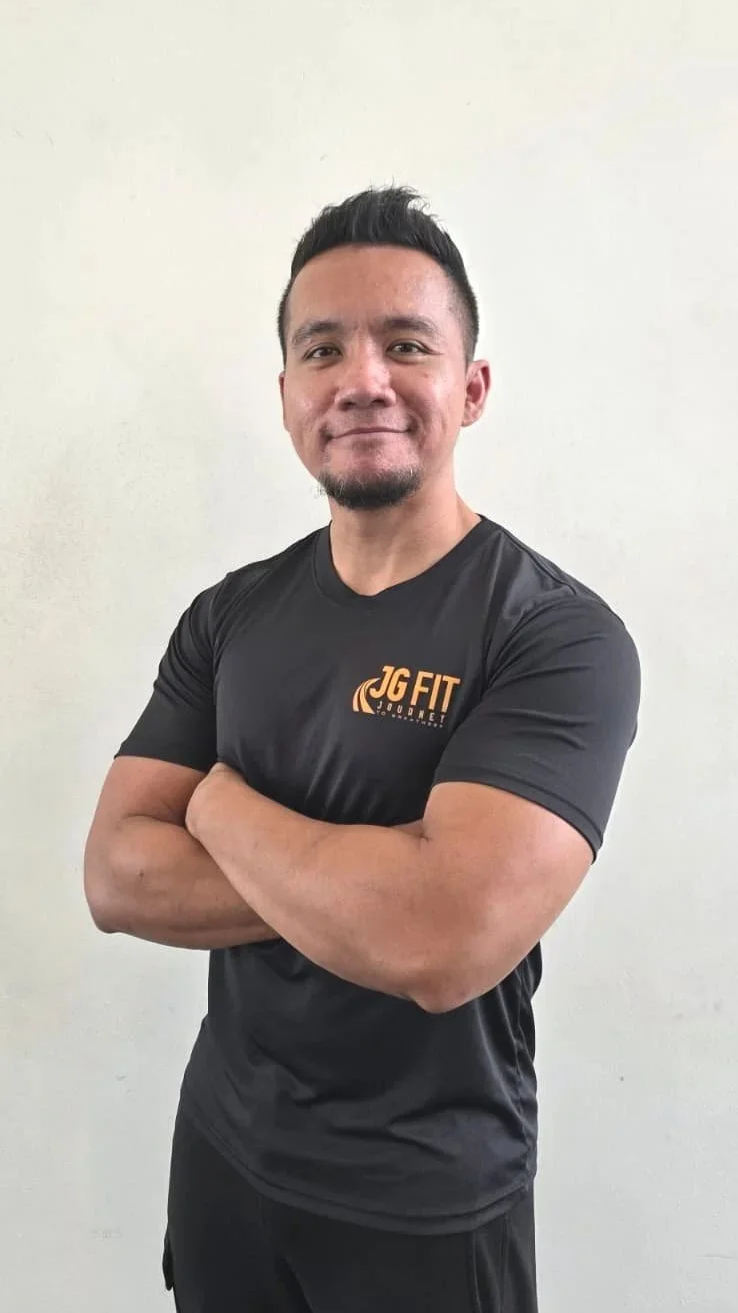 A man standing with arms crossed, wearing a black JG Fit T-shirt, smiling, against a plain light-colored background.