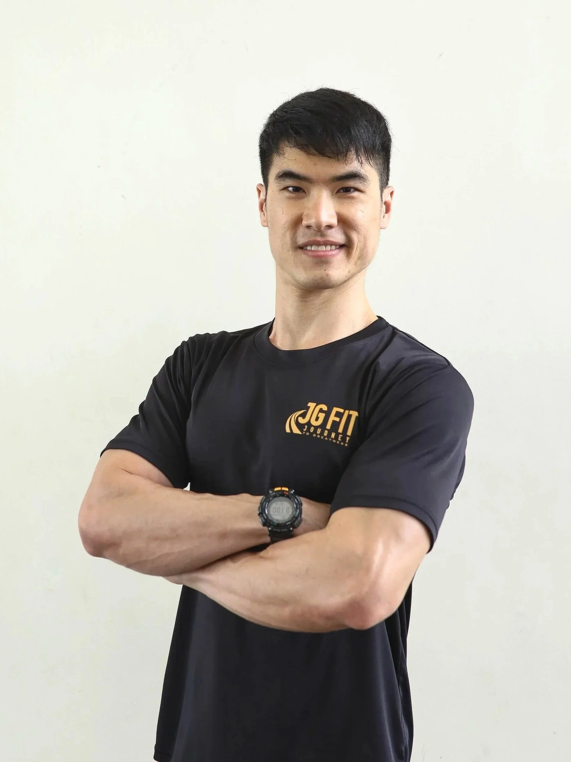 A young man with short black hair, wearing a black athletic T-shirt with the logo 'JG FIT JOURNEY' on it, standing with arms crossed against a plain light-colored wall.
