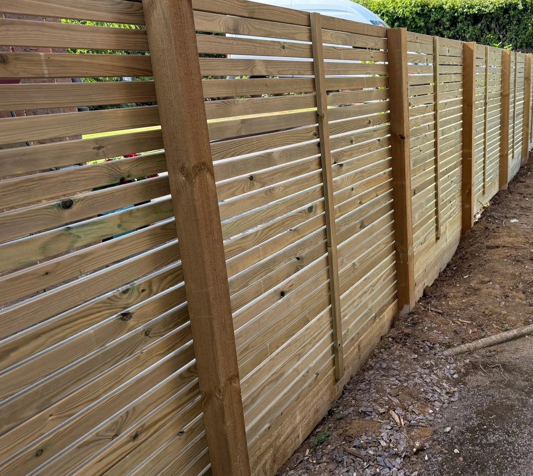 A newly constructed wooden outdoor privacy fence with horizontal slats and vertical support beams, with a dirt and gravel ground alongside.