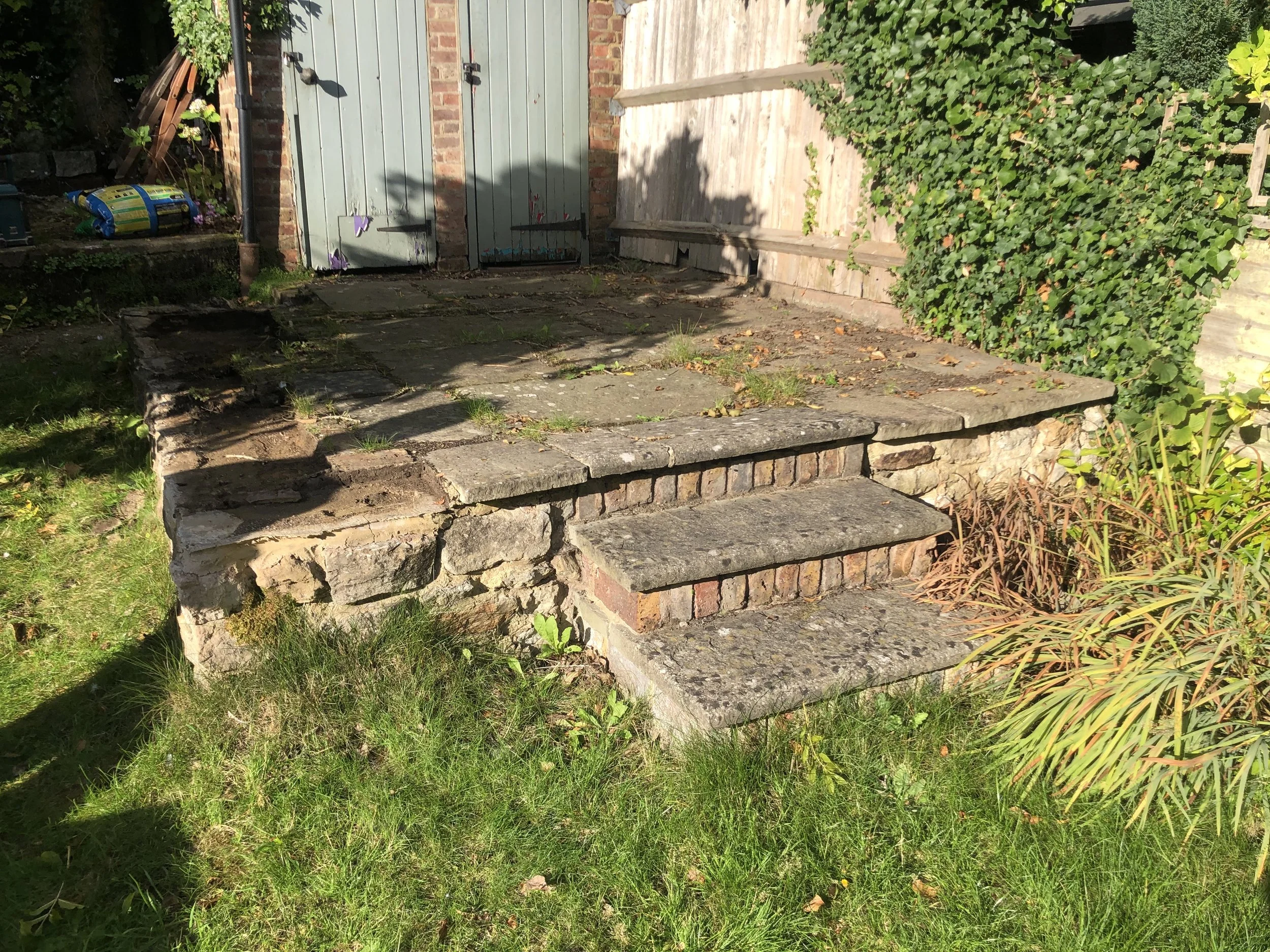 Stone steps leading up to a small elevated patio area with a wooden fence, green ivy, and a brick wall with light blue doors. There are some garden plants and grass in the foreground.