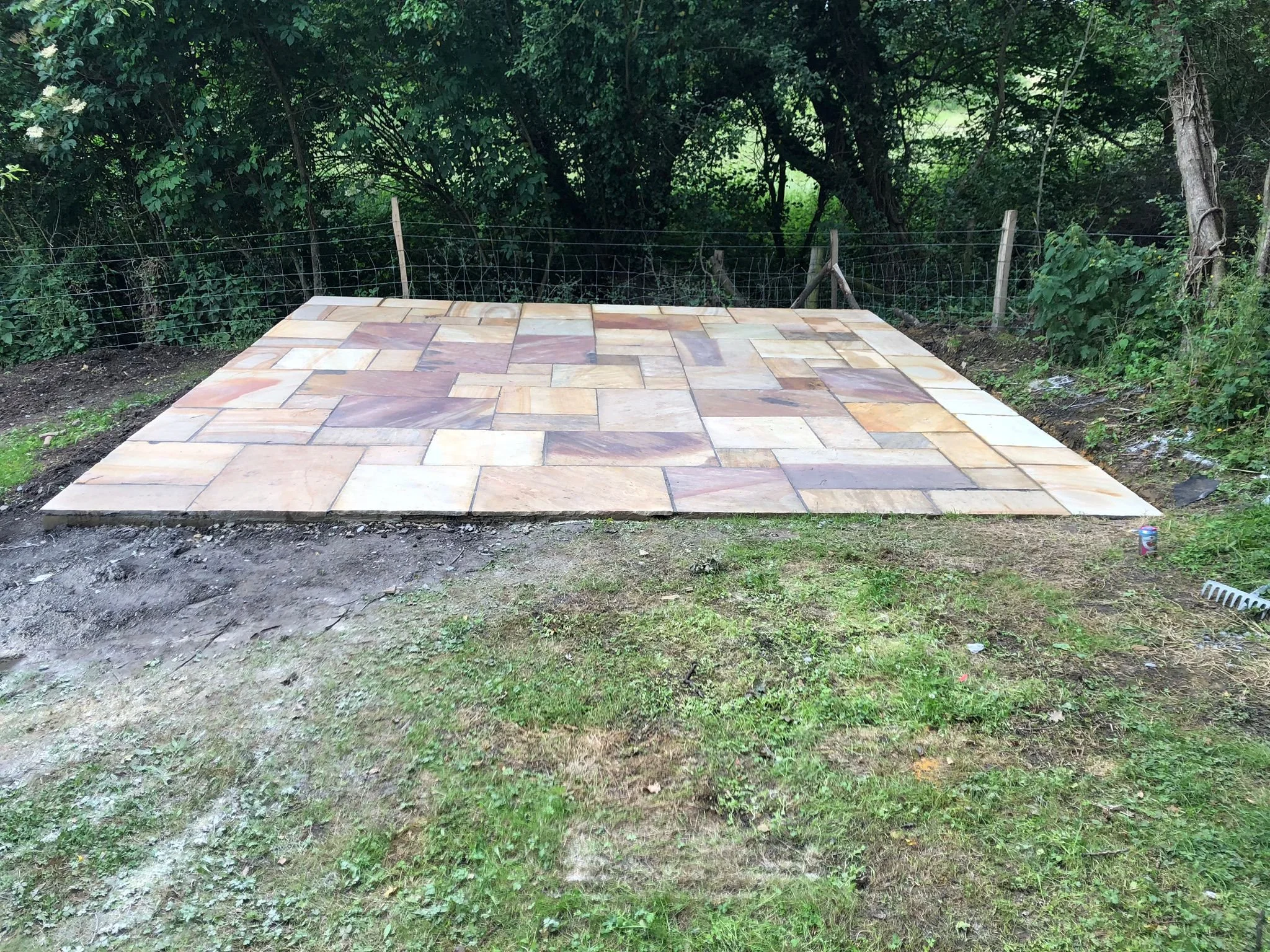 Freshly laid stone patio in a backyard with a wire fence, surrounded by green trees and grass.