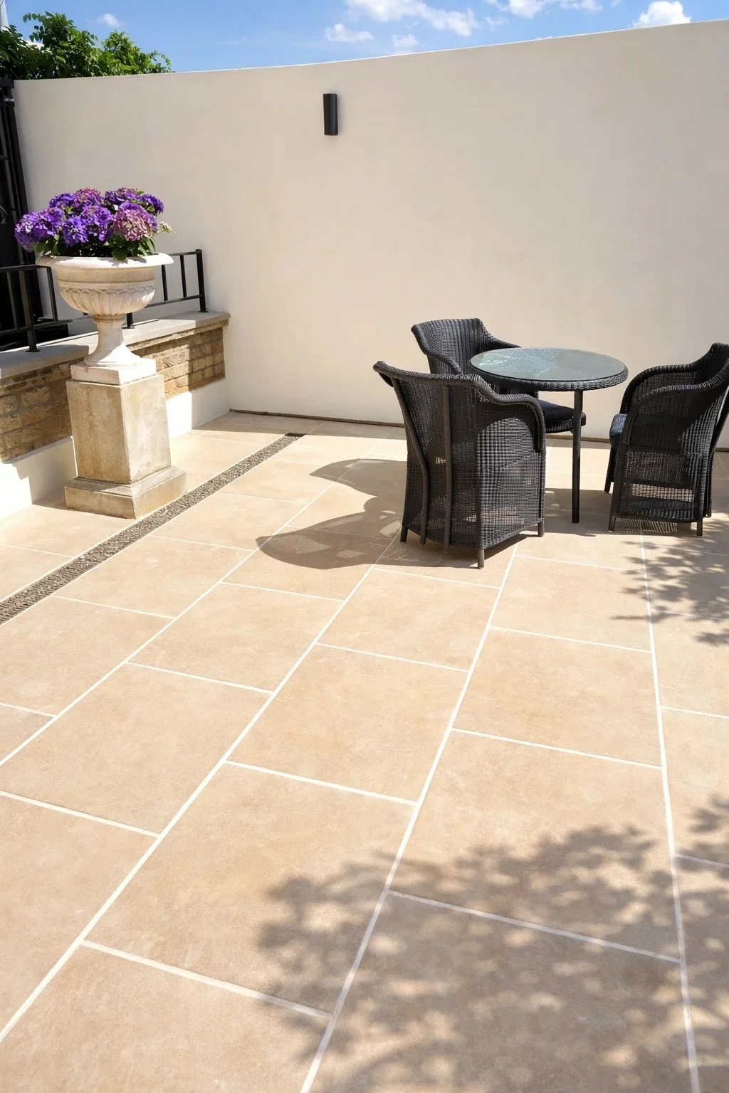 A patio with beige tiled flooring, a small round glass table surrounded by three black woven chairs, a large white planter with purple flowers, and a plain white wall with a black wall sconce, under a blue sky.