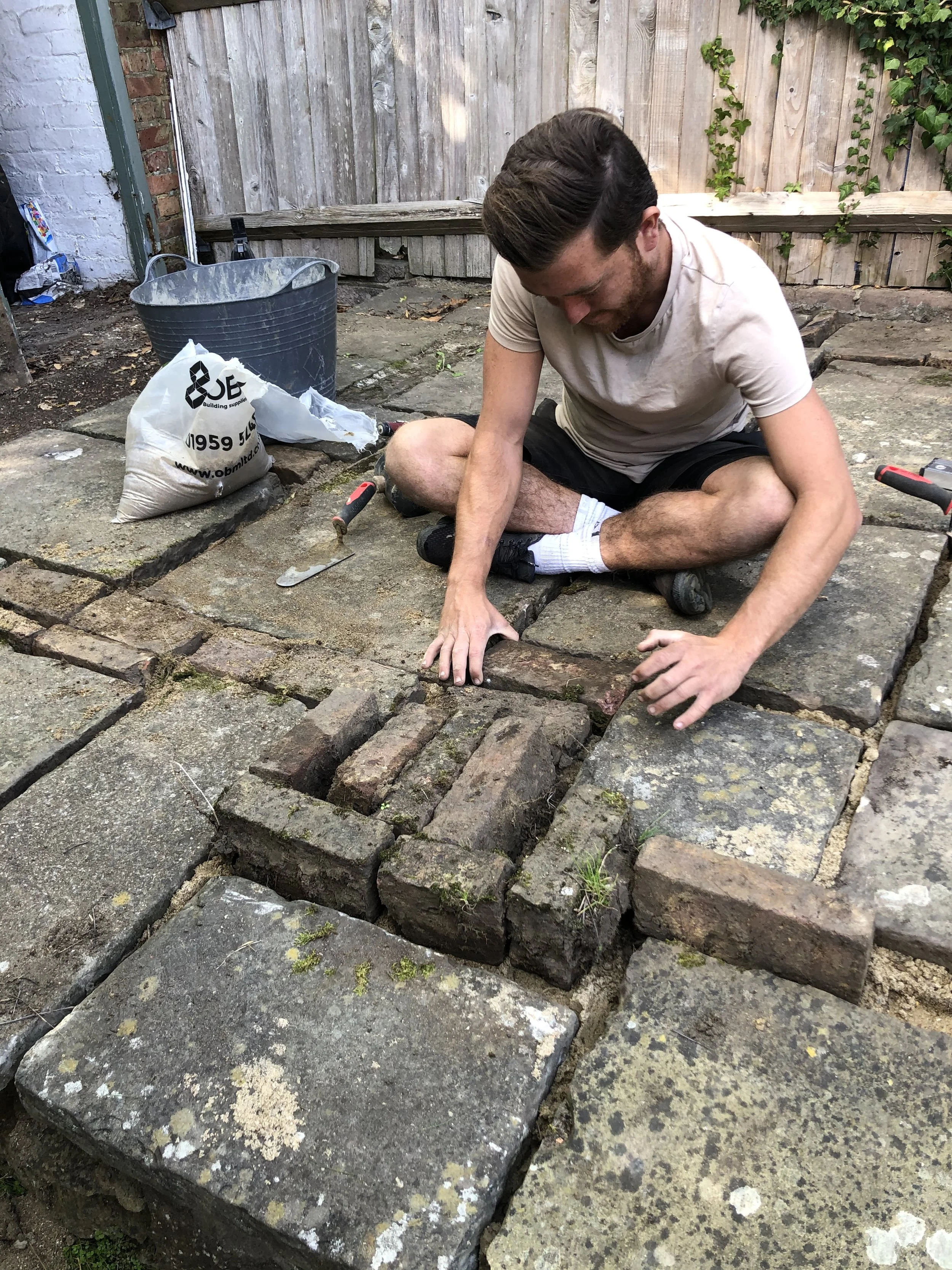 A man is sitting on the ground, laying brick pavers on a patio or walkway. He is surrounded by tools and materials, including a trowel, a bag of mortar, a bucket, and a few pavers.