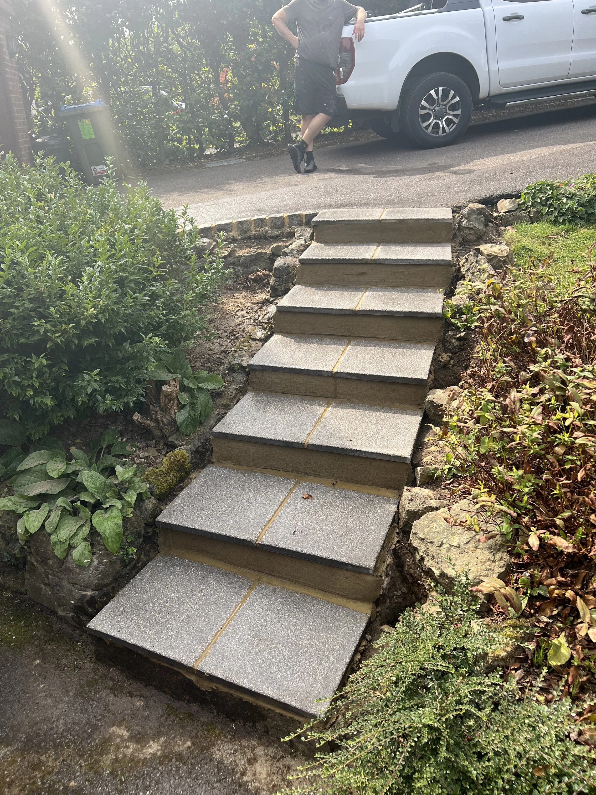 A set of eight concrete steps leading from a sidewalk to a driveway, with greenery and plants on either side, and a person leaning against a white pickup truck in the background.