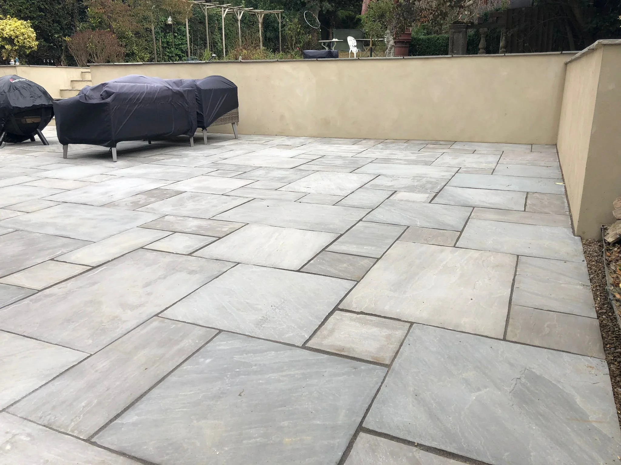 A spacious backyard patio with light gray stone tiles, two covered outdoor furniture pieces, and a beige stucco wall along the perimeter. In the background, there's a garden area with trees, shrubs, and patio furniture.