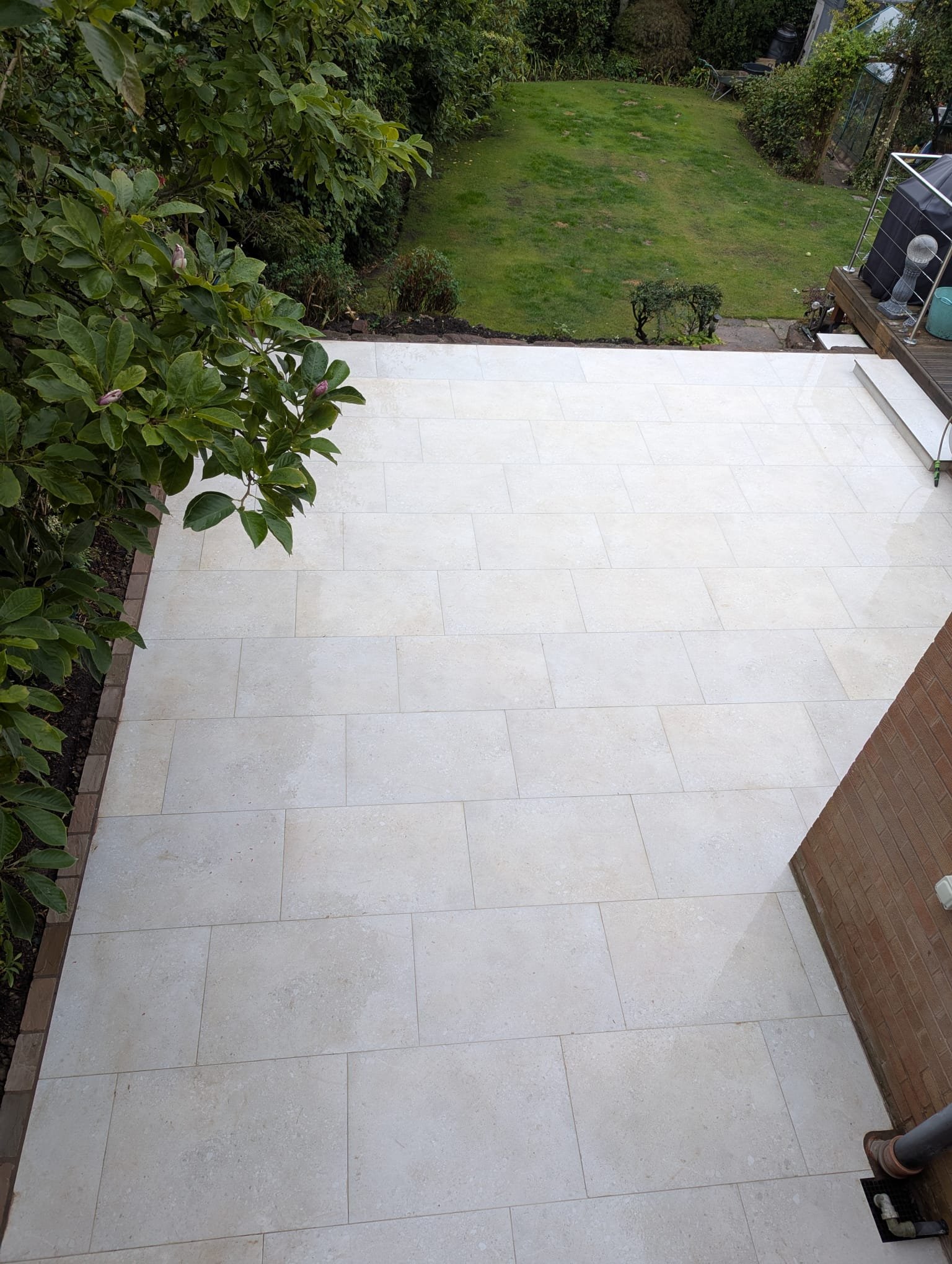View of a patio with large beige tiles, a garden with green grass and bushes, and a backyard with a lawn and trees.