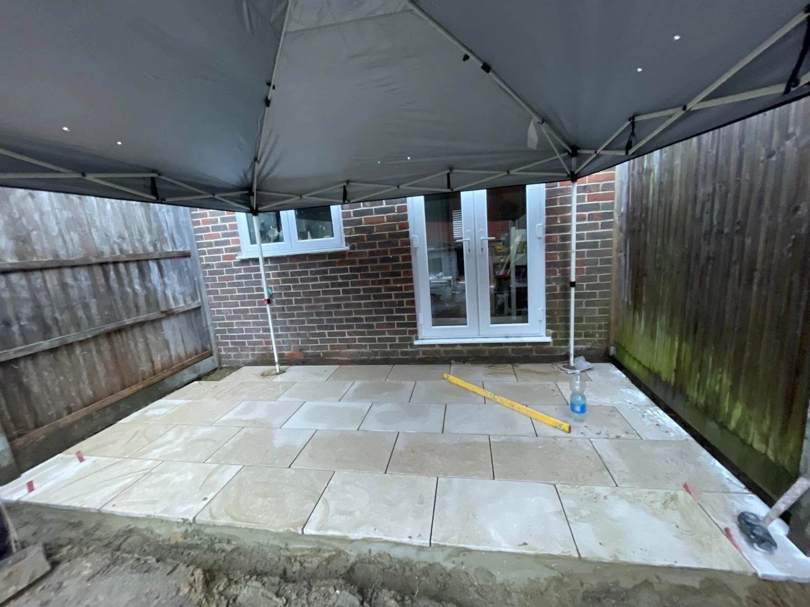 Backyard patio under construction with beige paving stones, a level tool, and a spray bottle, fenced by wooden panels with a brick house in the background.