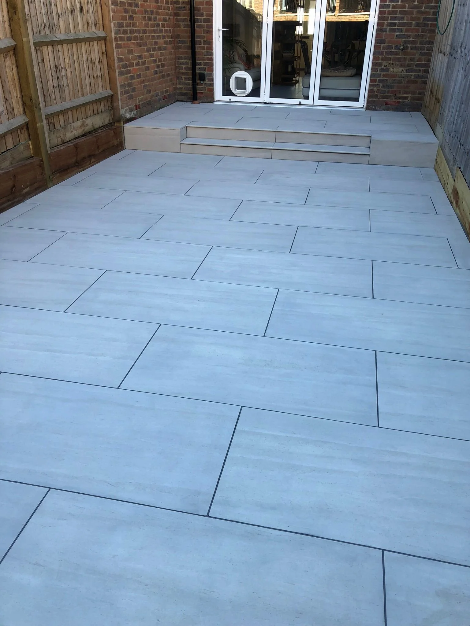 Backyard patio with large, light-colored ceramic tiles, wooden fencing, and a set of three small steps leading to a glass sliding door of a brick house.