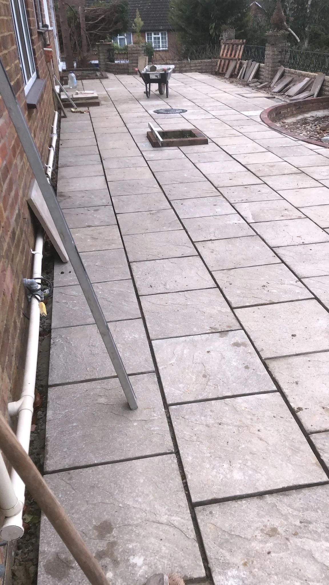 A backyard patio under construction with paving stones, a wheelbarrow, and tools. A section of the surface is unfinished.