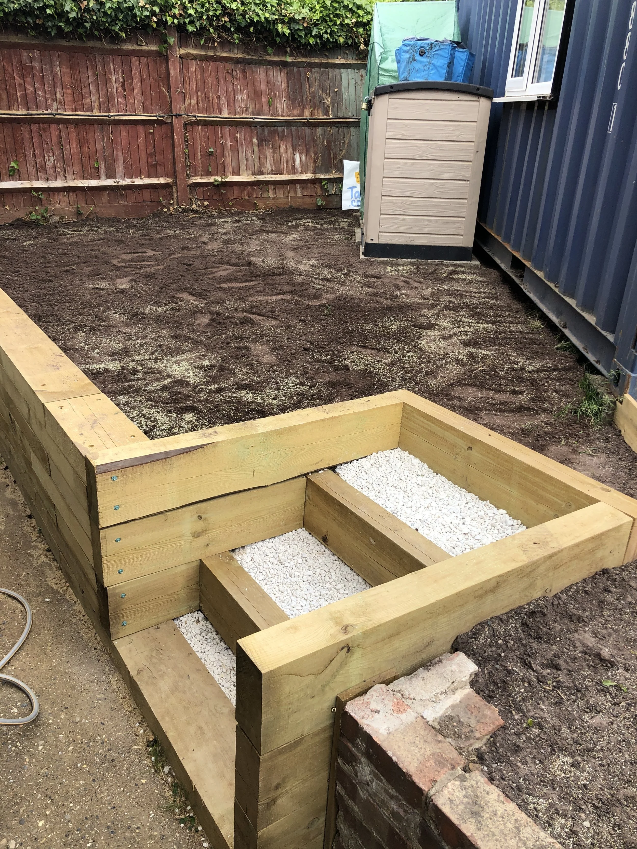 Wooden raised garden bed under construction, filled with white gravel, placed on a patch of bare soil next to a brick wall and a blue container, with a wooden fence in the background.
