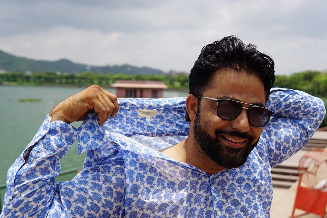 Man in sunglasses smiling and lifting a blue patterned shirt on a boat with water and green hills in the background.