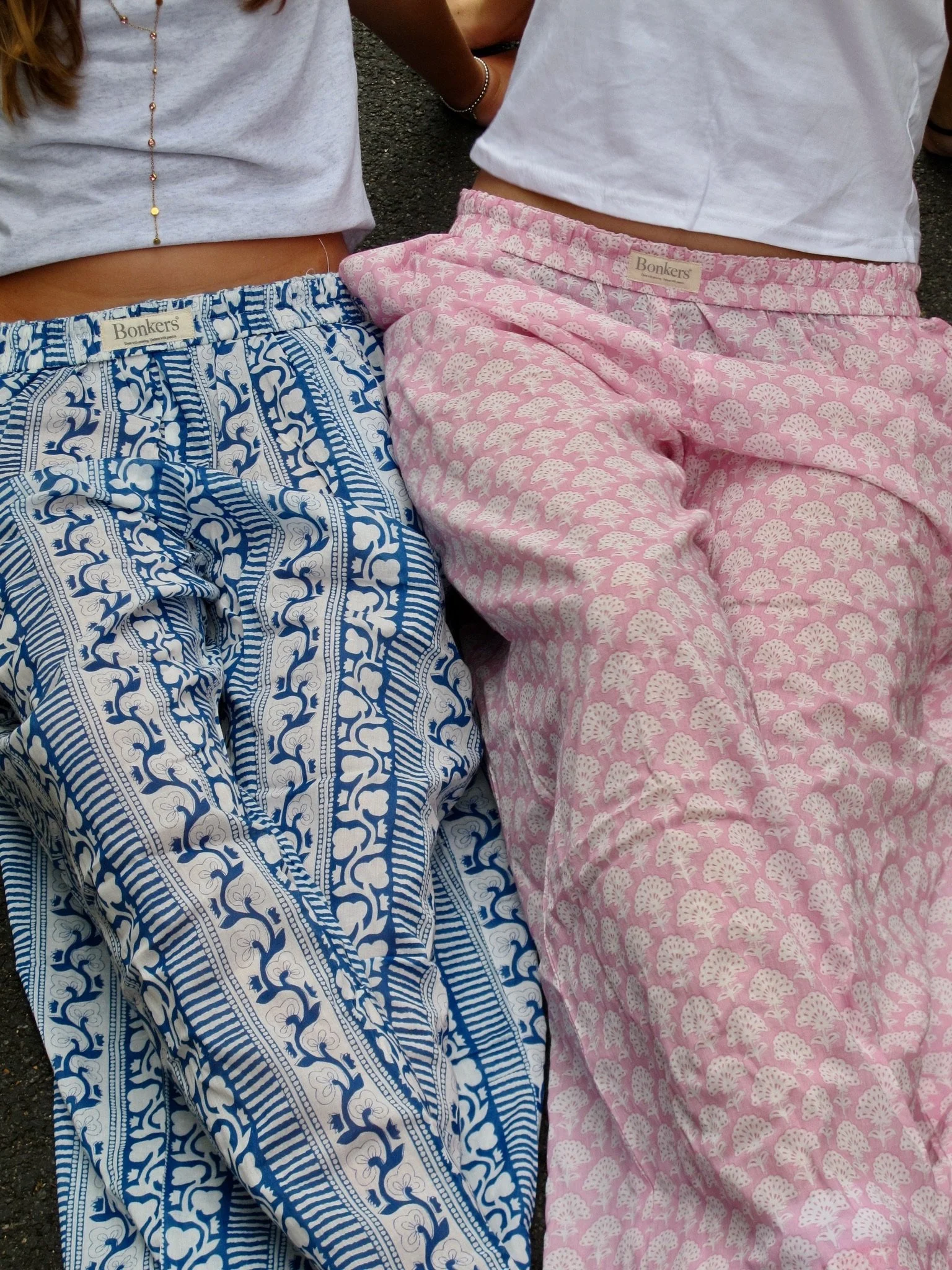 Close-up of two people wearing patterned pajama bottoms, one in blue and white, and the other in pink and white, with partial view of their torsos and legs, and a textured ground surface underneath.