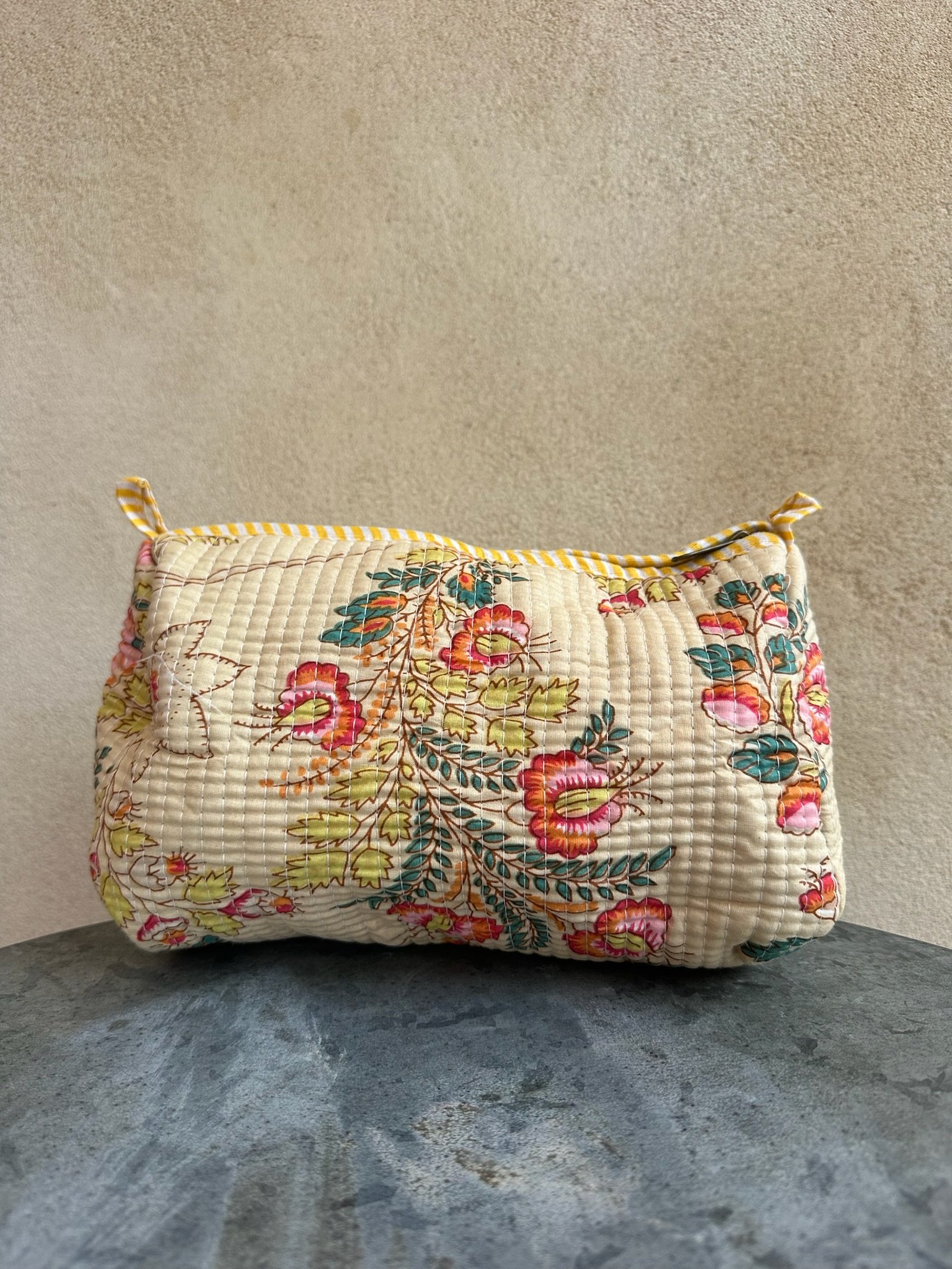 A floral quilted pillow with yellow striped trim resting on a concrete floor against a beige wall.