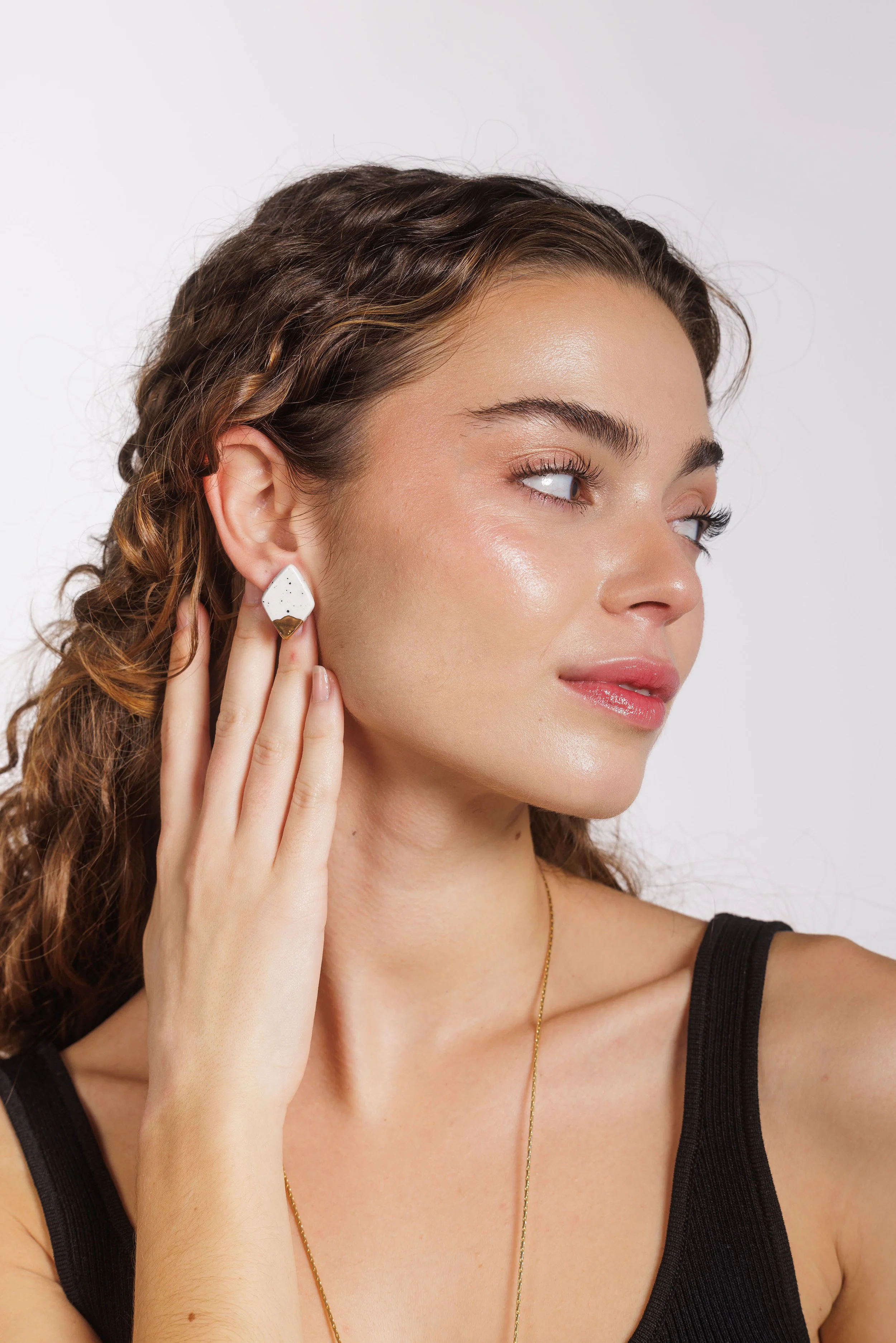 Retrato de una mujer joven con cabello rizado y piel radiante, usando aretes blancos y dorados, una camiseta negra y un collar dorado, tocándose la oreja izquierda con la mano derecha, en un fondo blanco.