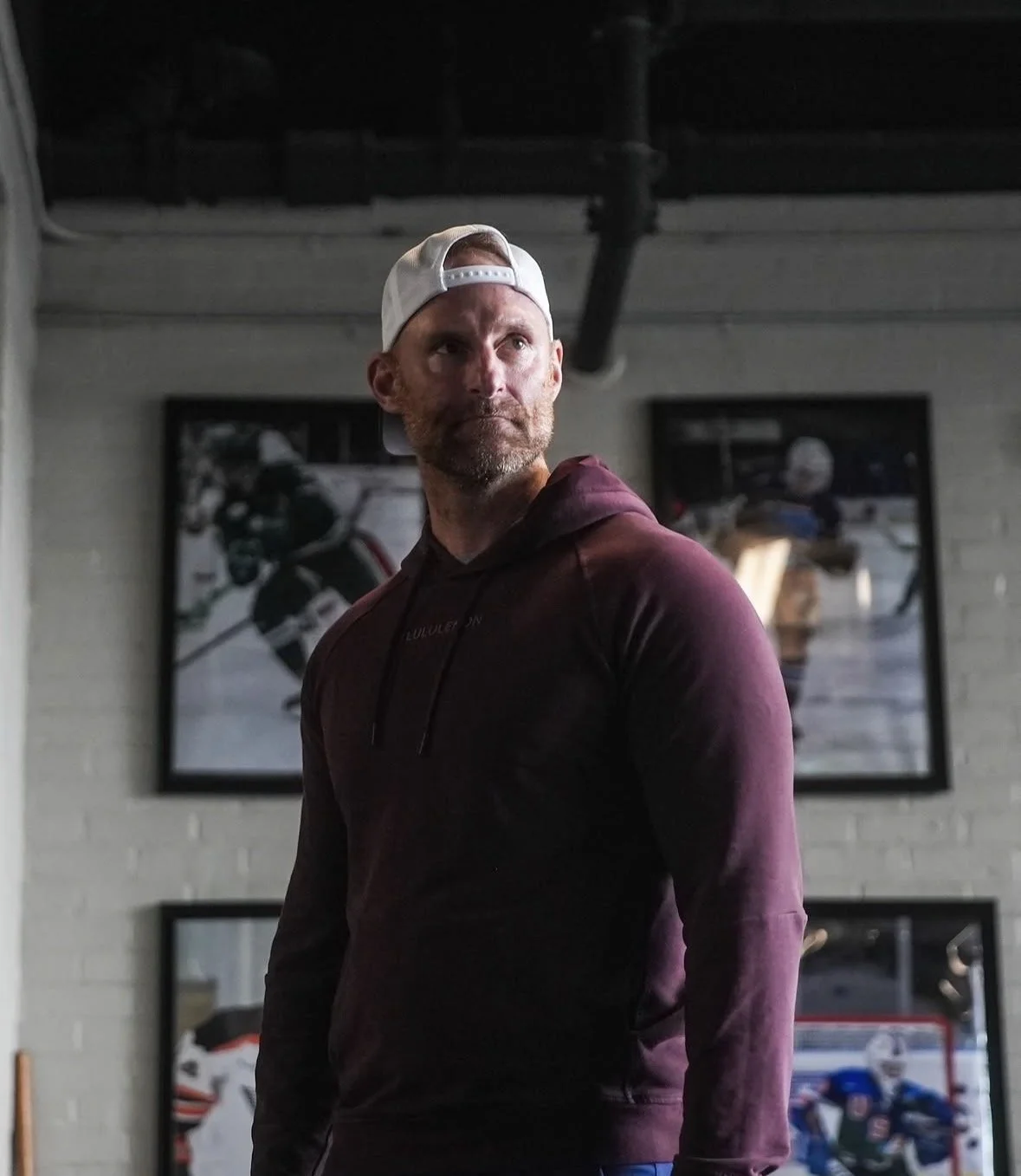 A man with a beard in a burgundy hoodie and a white backward cap standing in front of hockey posters.