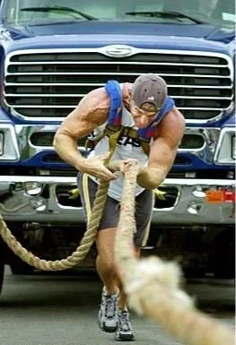 A man in workout attire is pulling a thick rope attached to a truck, with a dog holding onto the rope, in an outdoor setting.