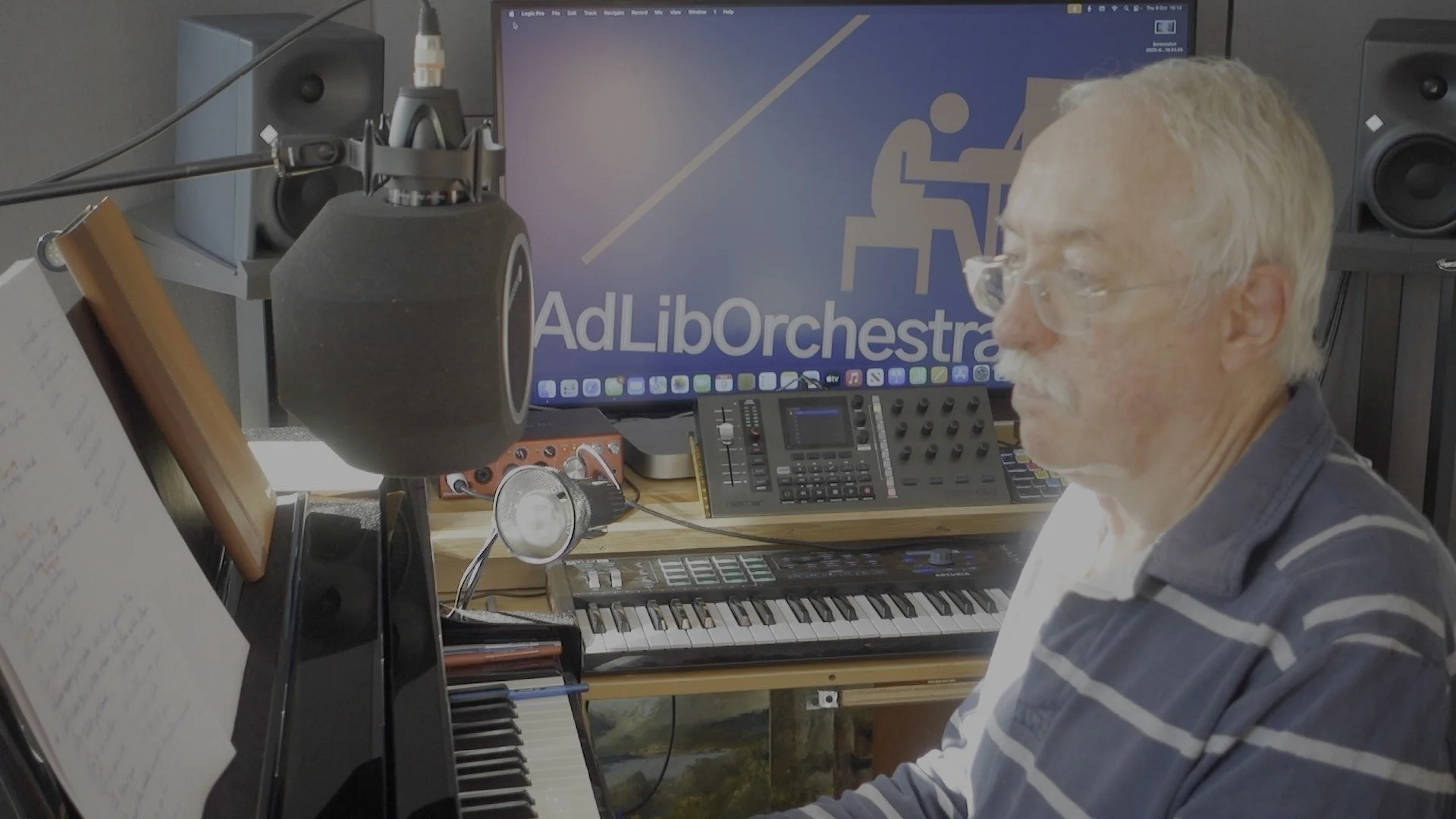 A man wearing glasses, seated at a music workstation with a keyboard, computer monitor with 'AdLibOrchestra' logo, studio speakers, and various audio equipment.