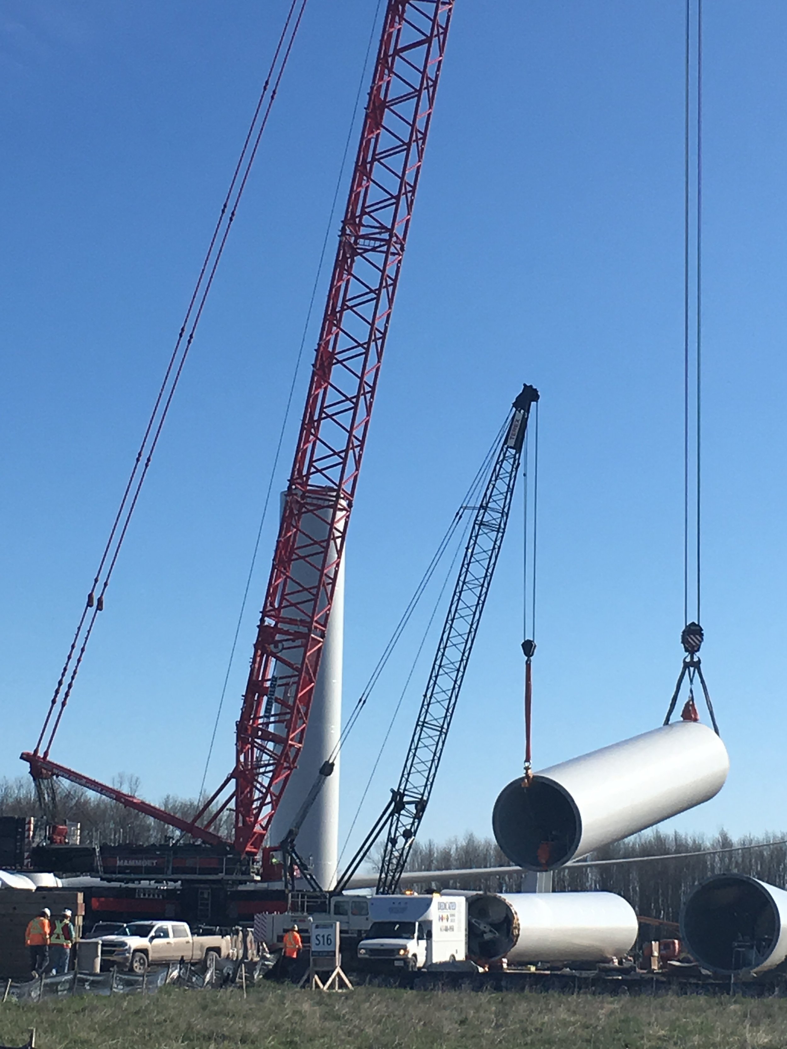 Construction site with large wind turbine blades being lifted by cranes, and workers in safety vests and helmets on the ground.