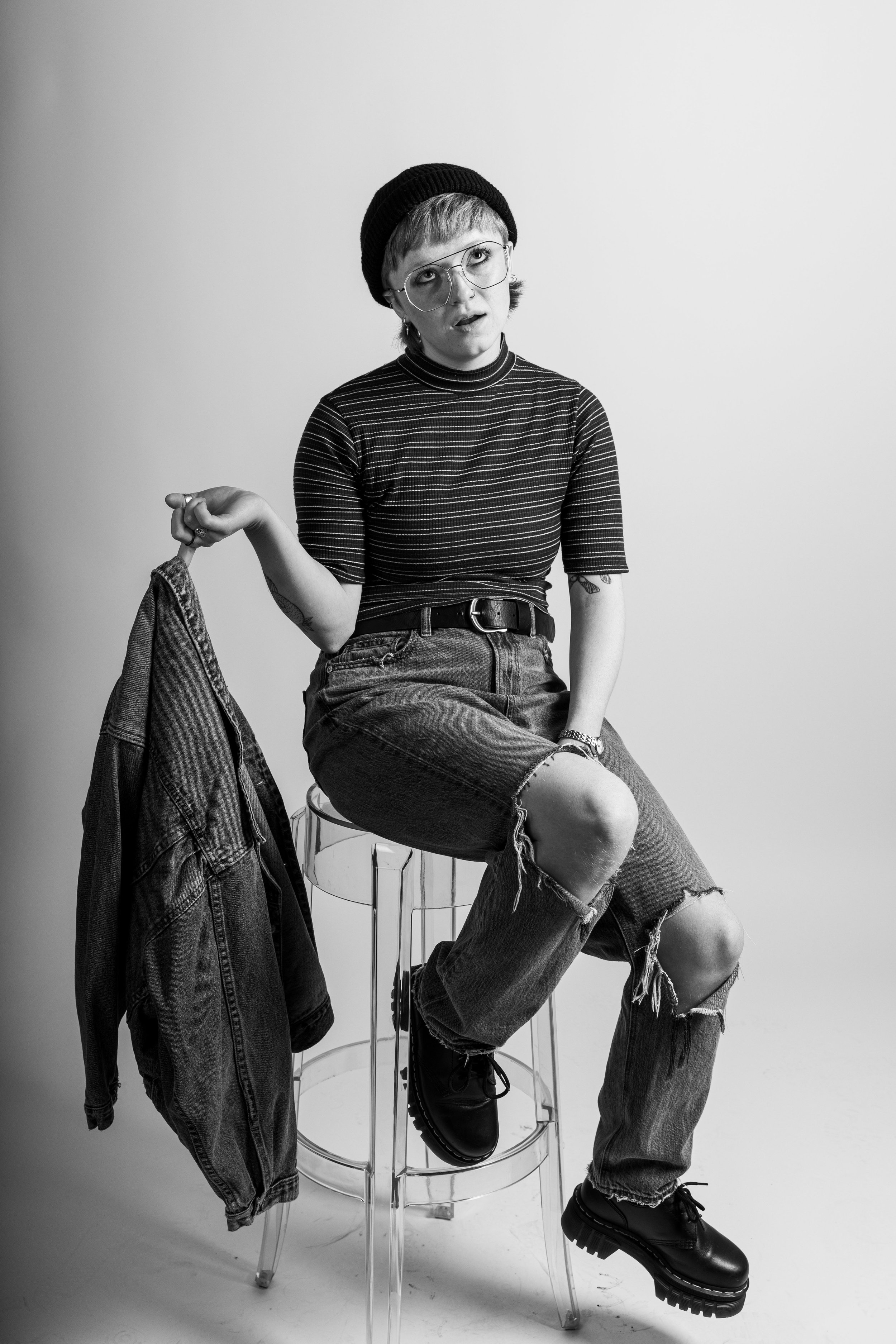 A black and white image of Finn wearing a black beanie, aviator glasses, a striped mock neck shirt, ripped jeans, and Dr. Marten boots. They're sitting on a plastic stool. They are holding a jean jacket by one finger. They are looking up.