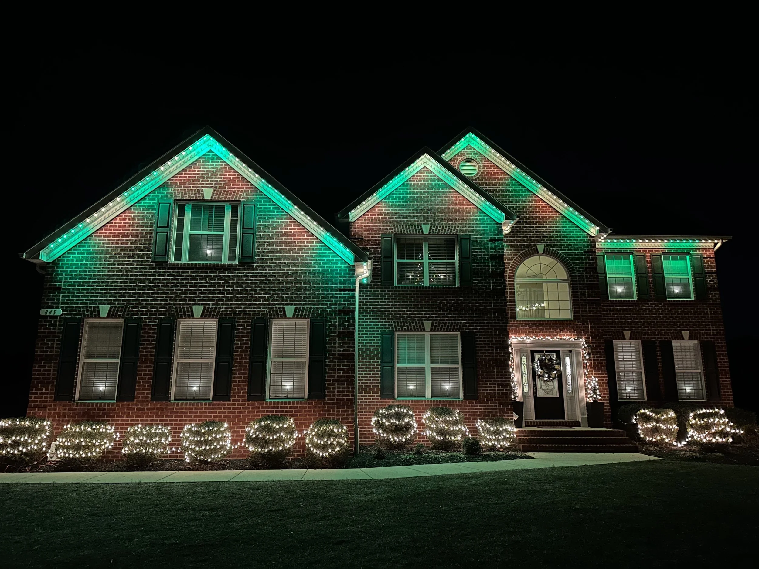 A two-story brick house decorated with green and white Christmas lights at night.