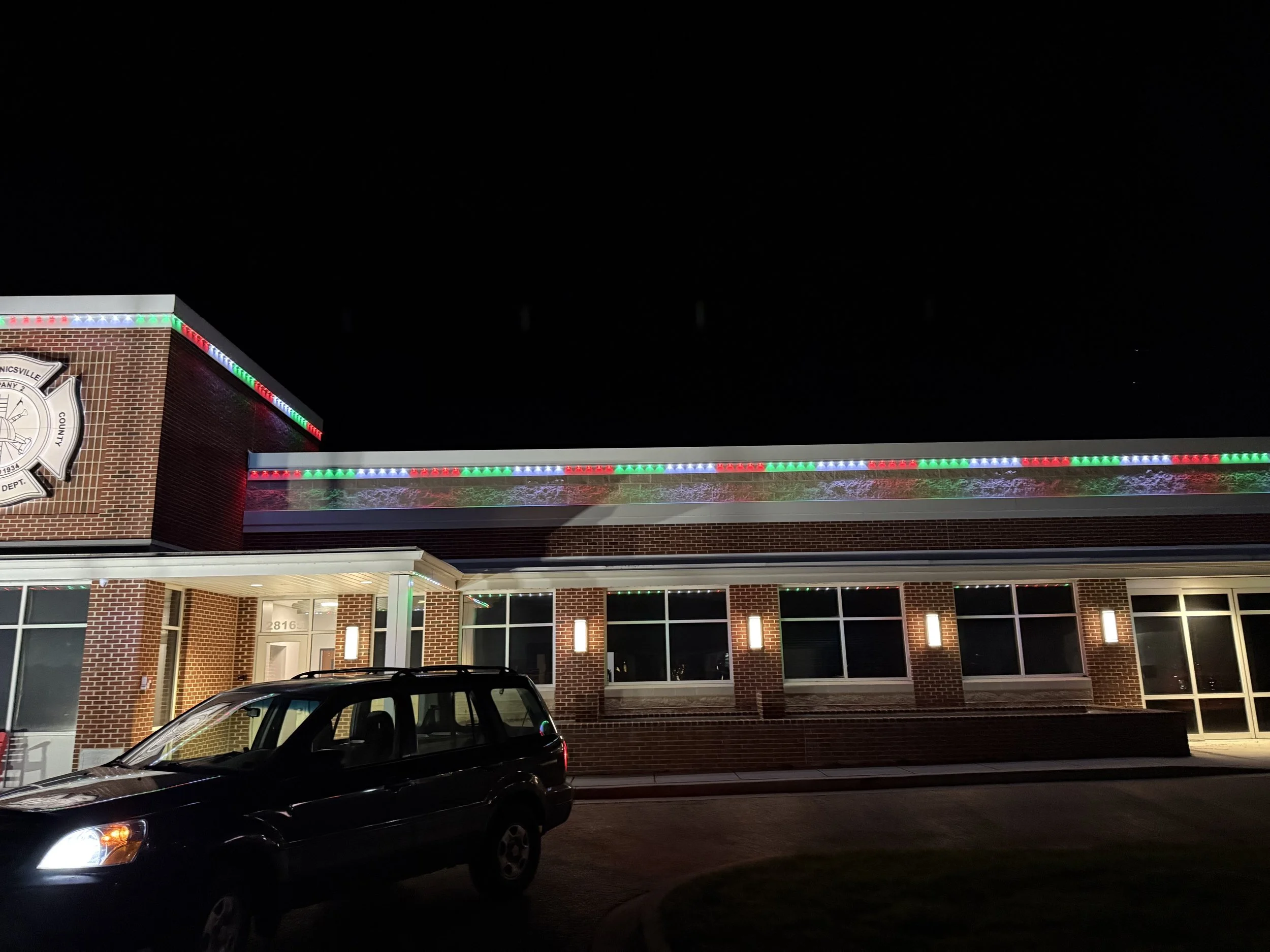 Night view of a brick building decorated with colorful Christmas lights along the roofline, a police badge emblem on the front, and a parked car in the foreground.