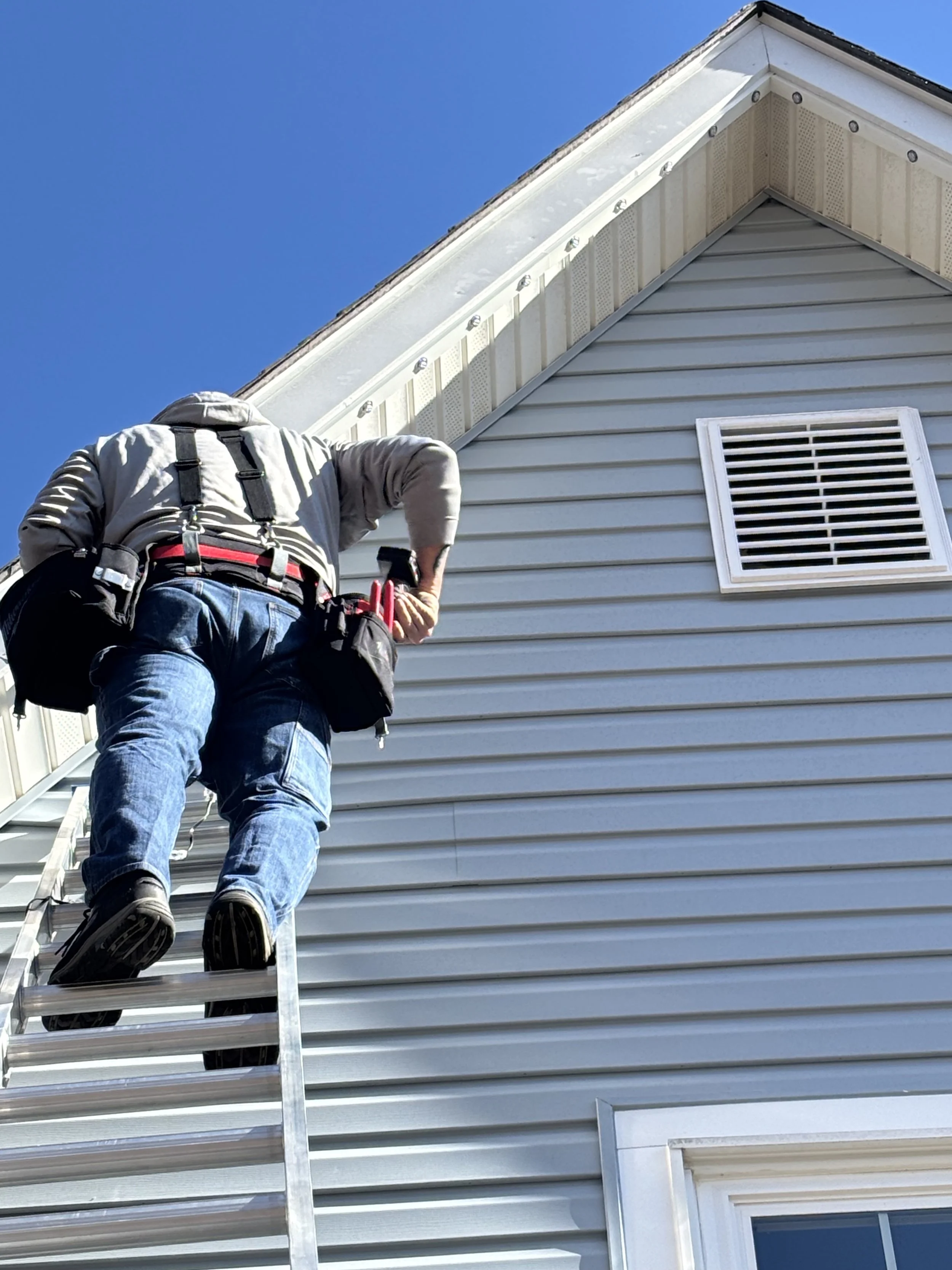 Worker climbing a ladder to clean the exterior siding of a house on a clear, sunny day.