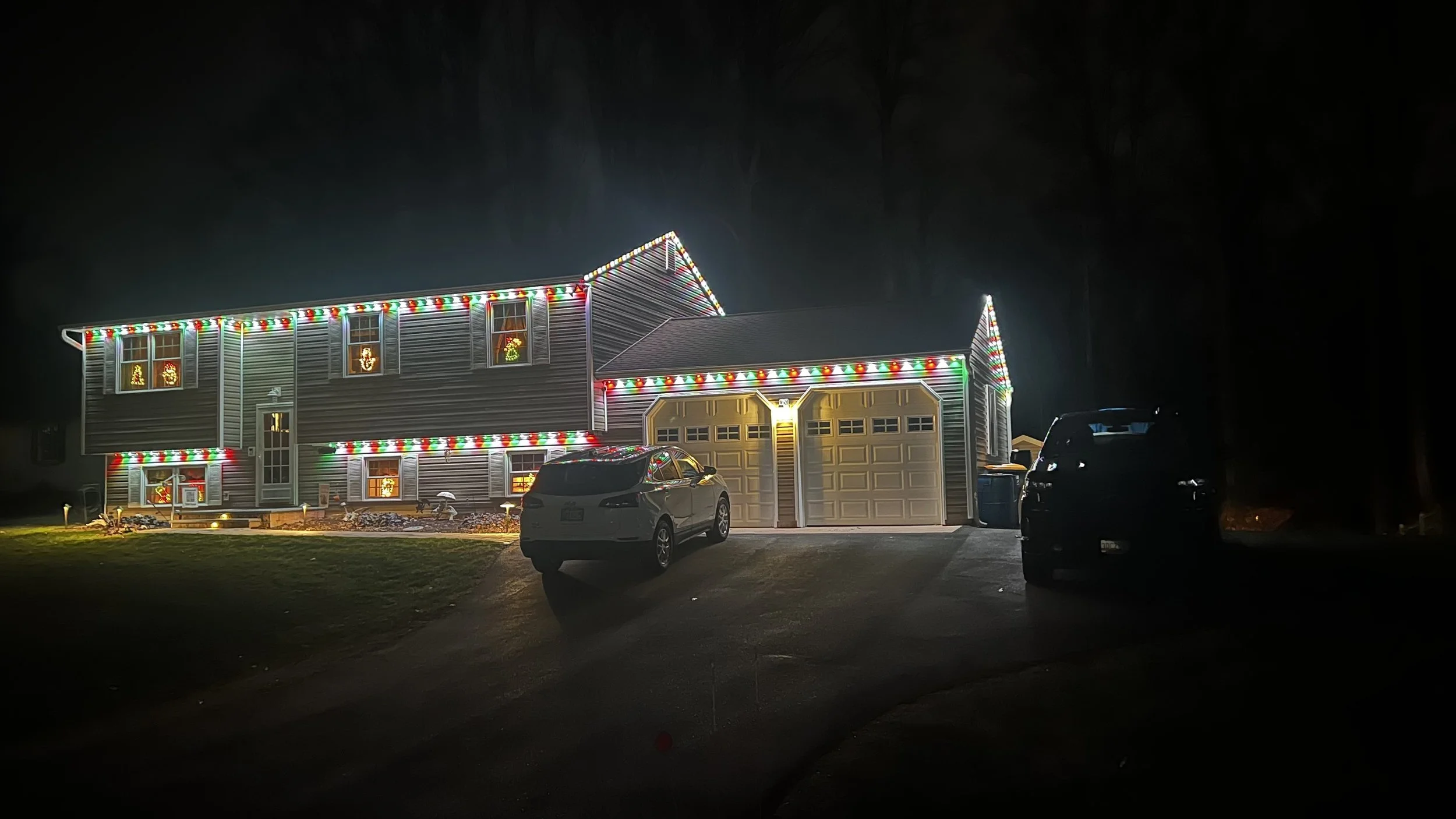 A house decorated with colorful Christmas lights at night, with cars parked in the driveway and illuminated windows.