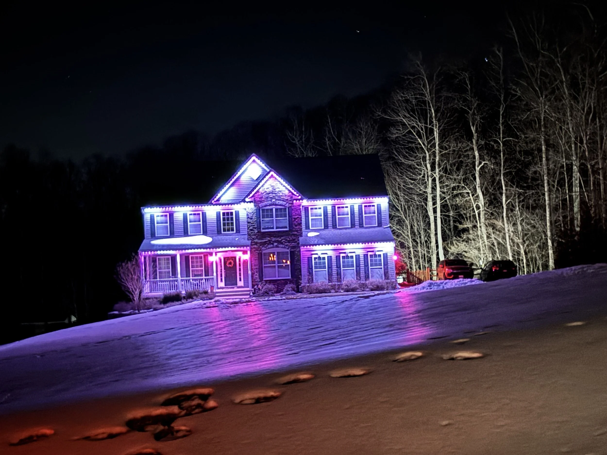 A house decorated with colorful Christmas lights during winter at night, with snow on the ground and footprints leading up to the house.