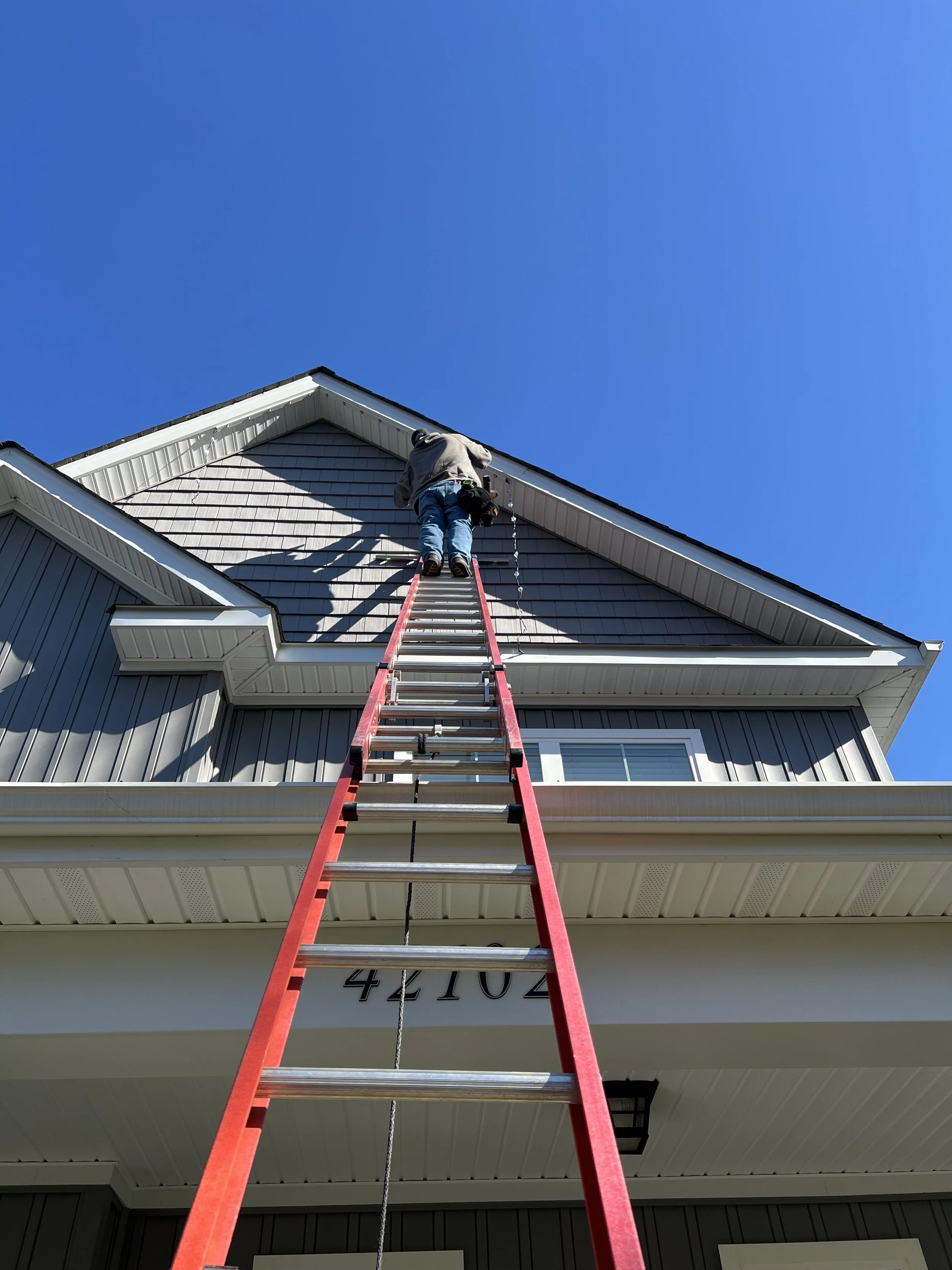 A person climbing a tall red ladder to the roof of a modern house with gray siding and white trim on a clear, sunny day.
