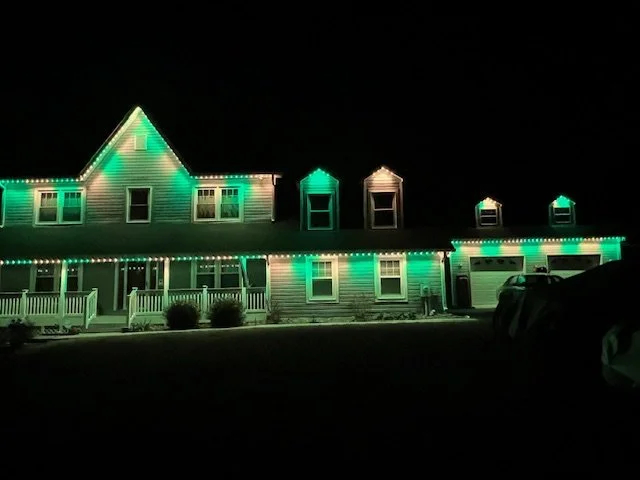 A large two-story house decorated with green and white Christmas lights at night.