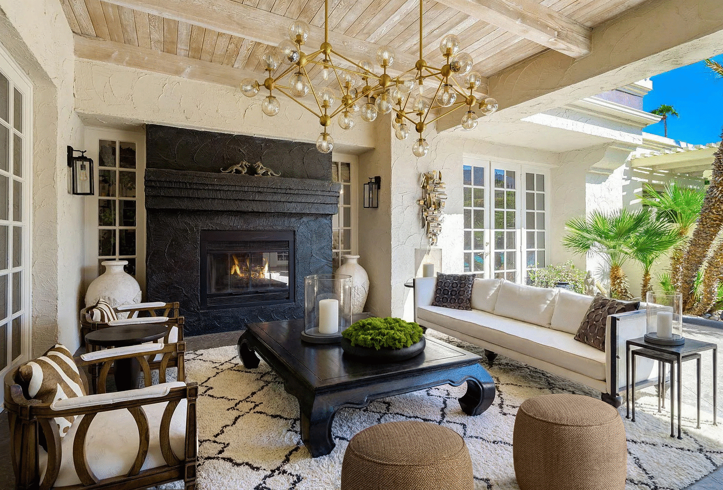 Palm Springs covered patio with brass chandelier, carved wood furniture, outdoor fireplace, French doors, palm trees, and indoor-outdoor desert entertaining space.