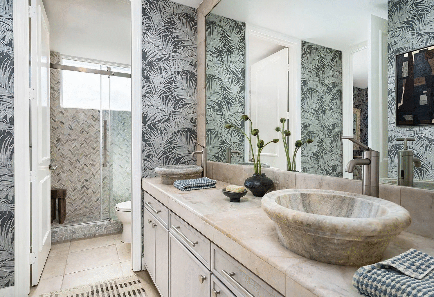 Palm Springs guest bathroom featuring stone vessel sinks, palm-pattern wallpaper, herringbone tile shower, and organic modern desert materials.