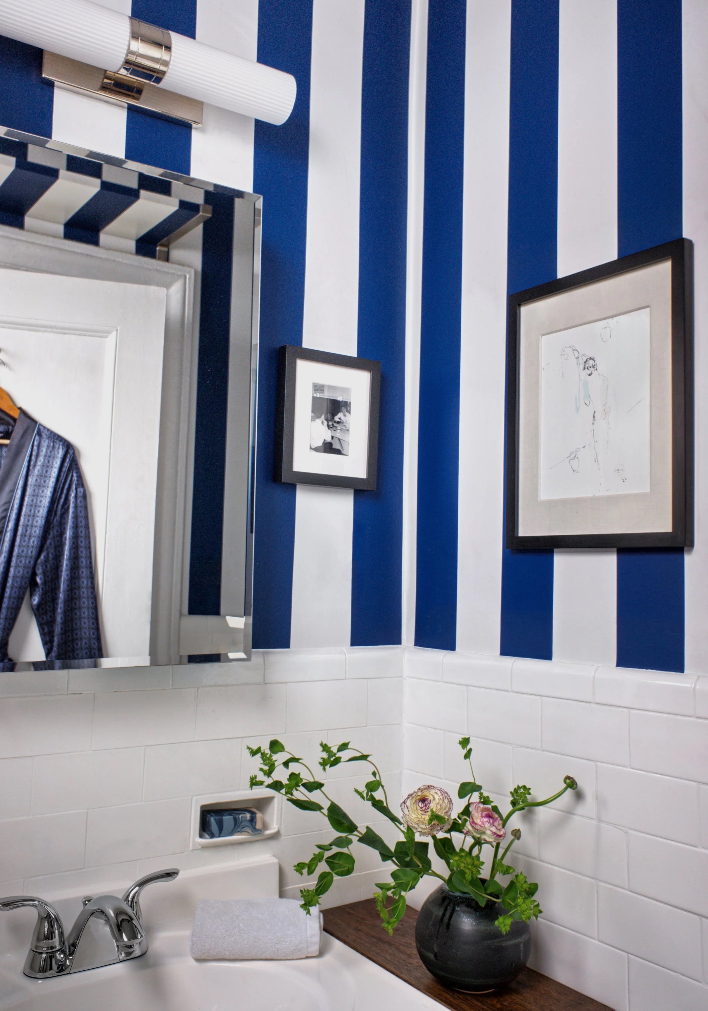 Manhattan bathroom with bold blue and white striped wallpaper, framed artwork, chrome fixtures, and styled floral arrangement in Hell’s Kitchen apartment.