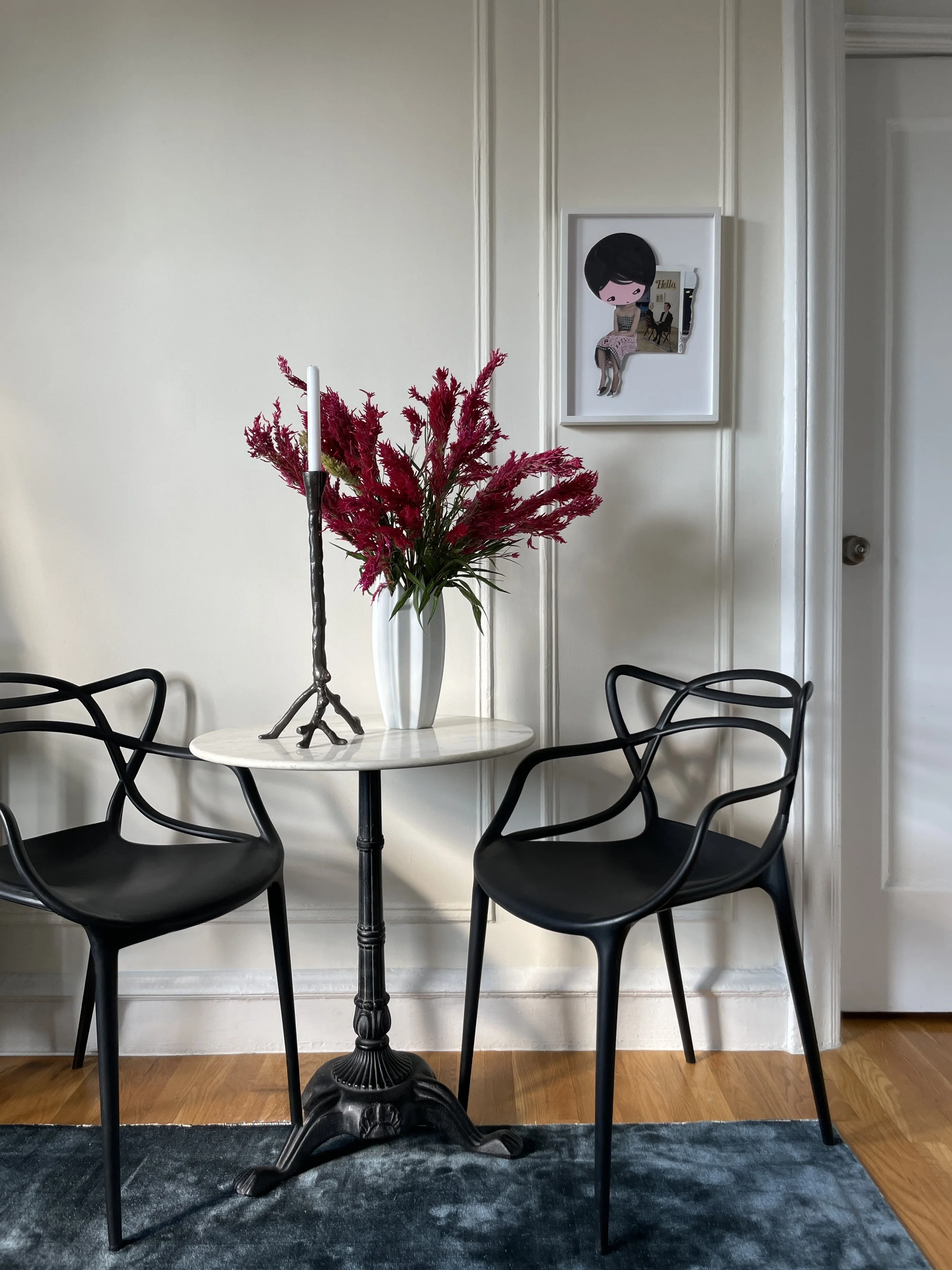 Small Parisian-inspired dining nook in Hell’s Kitchen apartment featuring round marble table, sculptural black Kartell Masters chairs, tall candlestick, red floral arrangement, and classic wall moldings in a New York prewar rental.