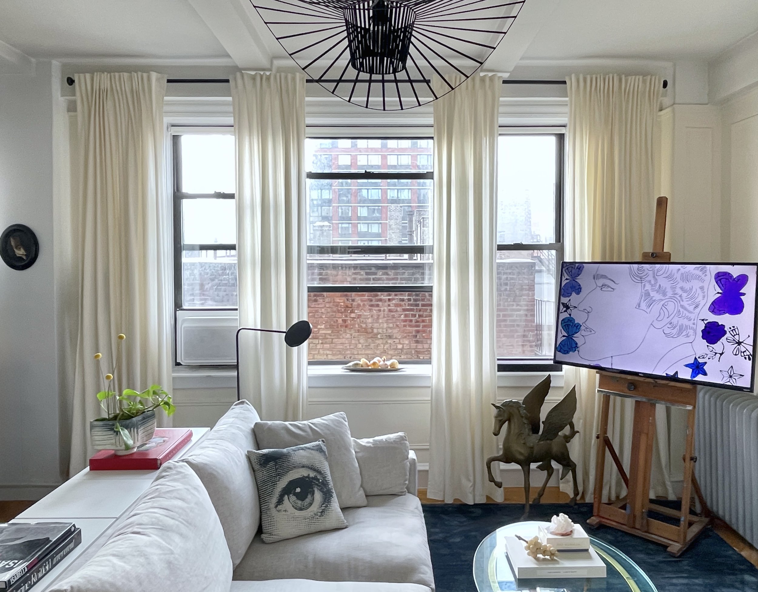 Hell’s Kitchen living room with retrofitted television mounted on vintage artist easel, tall ivory drapery panels, sculptural black ceiling light, Manhattan skyline views, and layered collected decor in a curated New York rental apartment.