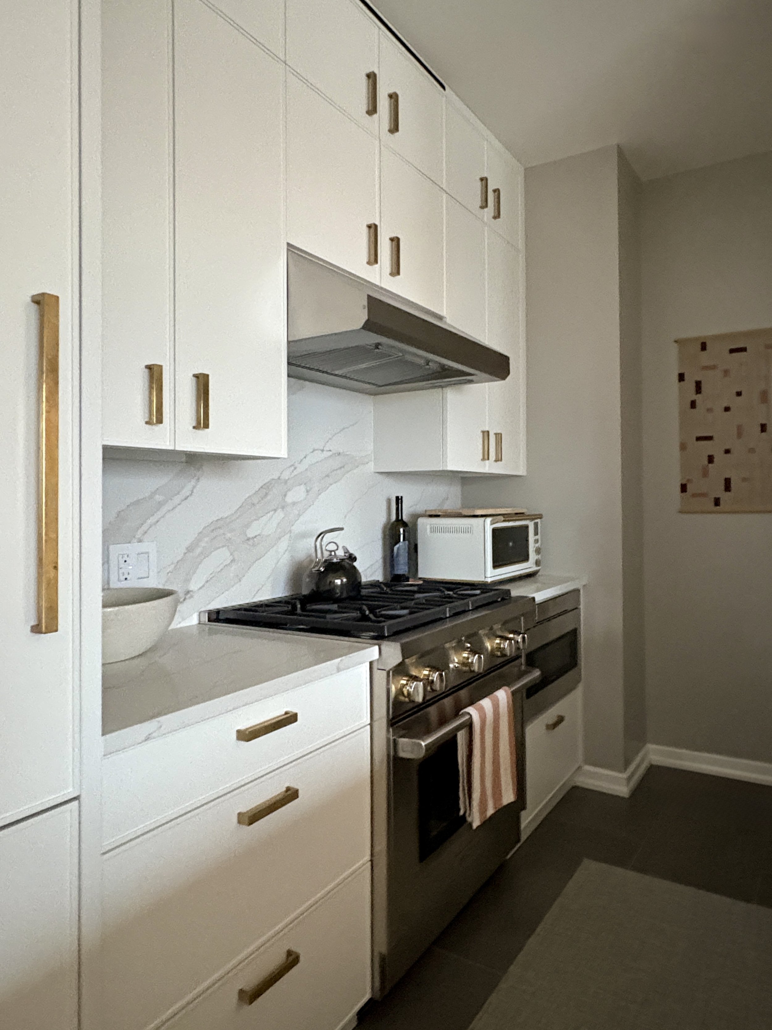 Custom Upper West Side kitchen featuring modern shaker-style white cabinetry, burnished brass Katonah hardware, marble backsplash with dramatic veining, stainless steel range, and tailored storage solutions in a full apartment renovation.