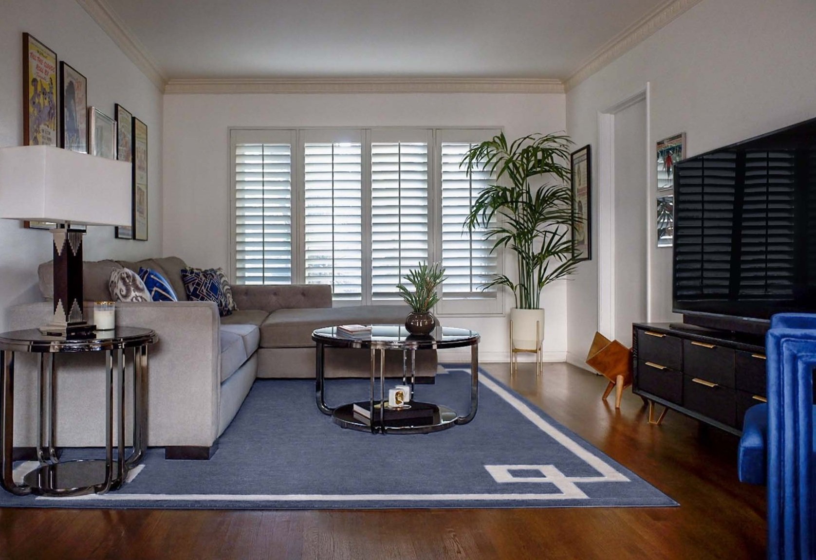 West Hollywood living room with gray sectional sofa, black nickel and glass coffee table, navy Greek key area rug, mid-century wood credenza, and layered vintage film poster collection creating Hollywood Regency meets Art Deco design.