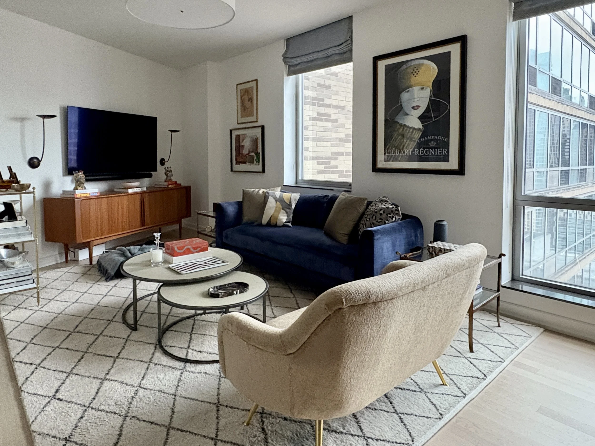 Upper West Side living room with deep navy velvet sofa, curved cream bouclé accent chair, mid-century wood credenza, layered neutral rug, curated art collection, and large Manhattan windows in a refined modern renovation.