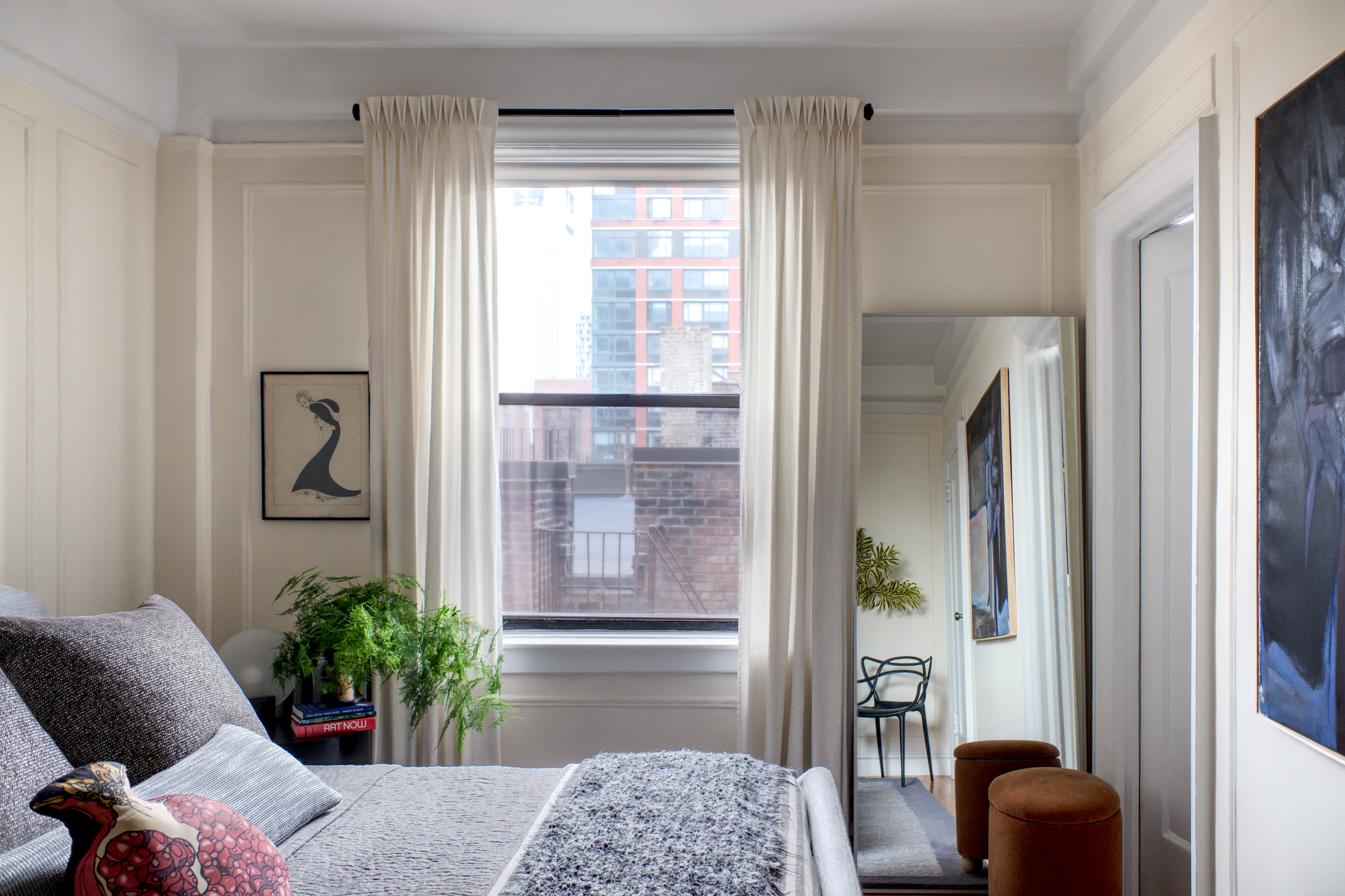 Hell’s Kitchen primary bedroom with custom ivory pleated drapery, tall New York windows, layered neutral bedding, oversized leaning mirror, cognac velvet ottomans, vintage artwork, and Parisian-inspired styling in a curated Manhattan rental.