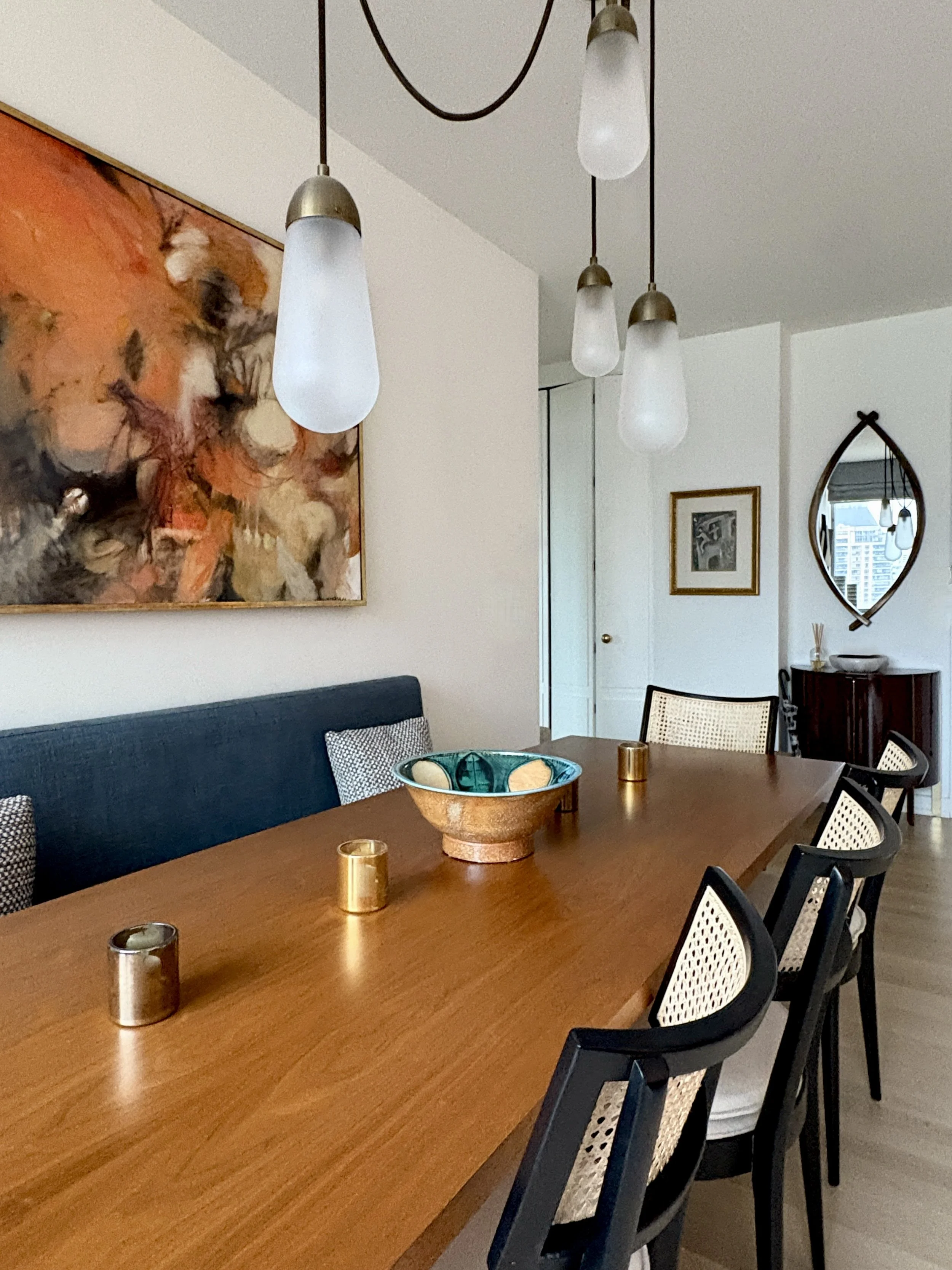 Upper West Side apartment dining room featuring Apparatus Larita pendant light cascading over a walnut dining table, cane dining chairs, abstract artwork, layered brass accents and skyline views in a full gut renovation.