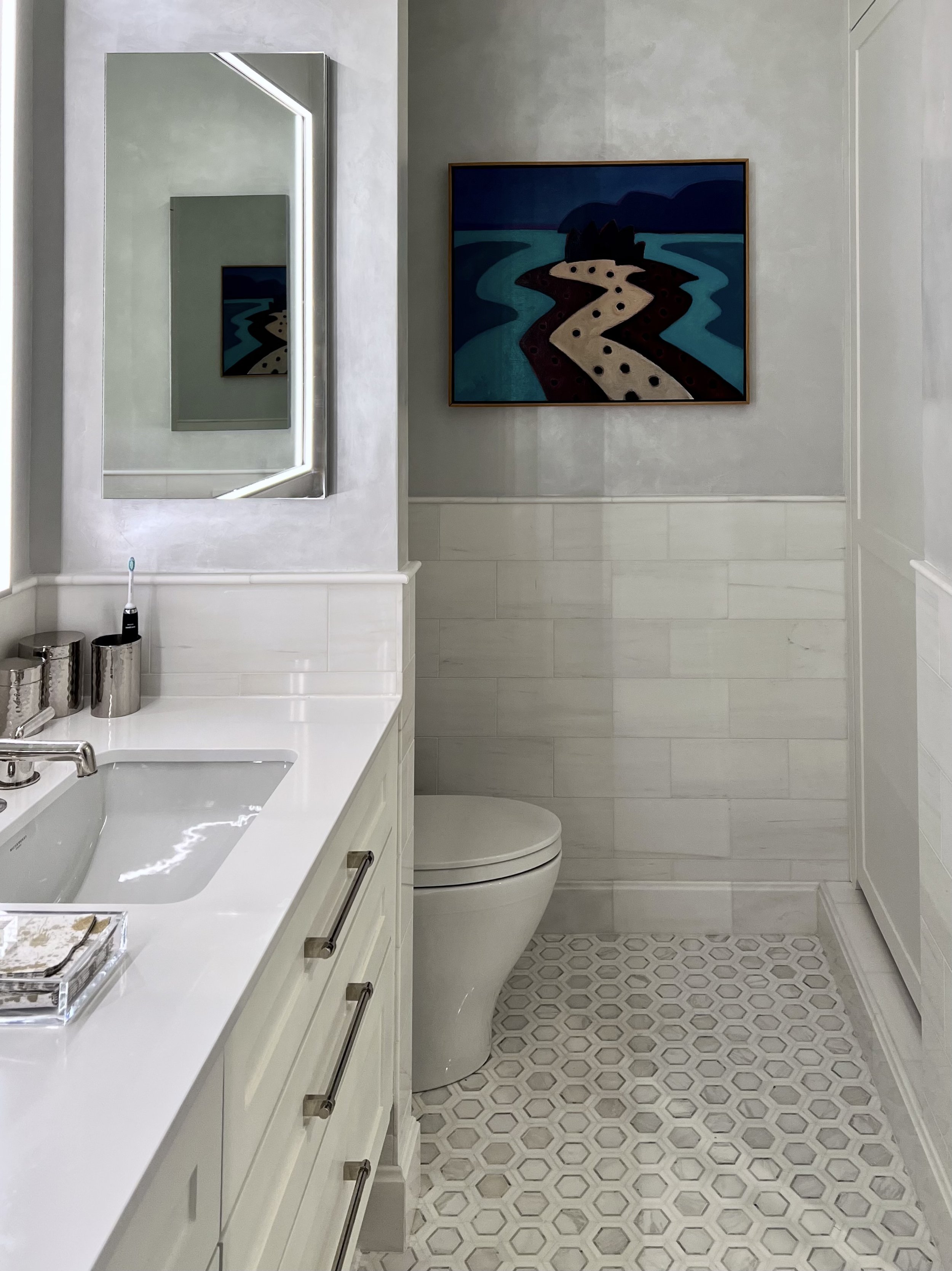 Upper West Side modern bathroom with marble hex tile flooring, white vanity cabinetry, integrated sink, textured plaster walls, framed contemporary artwork, and sleek illuminated mirror in a luxury New York renovation.