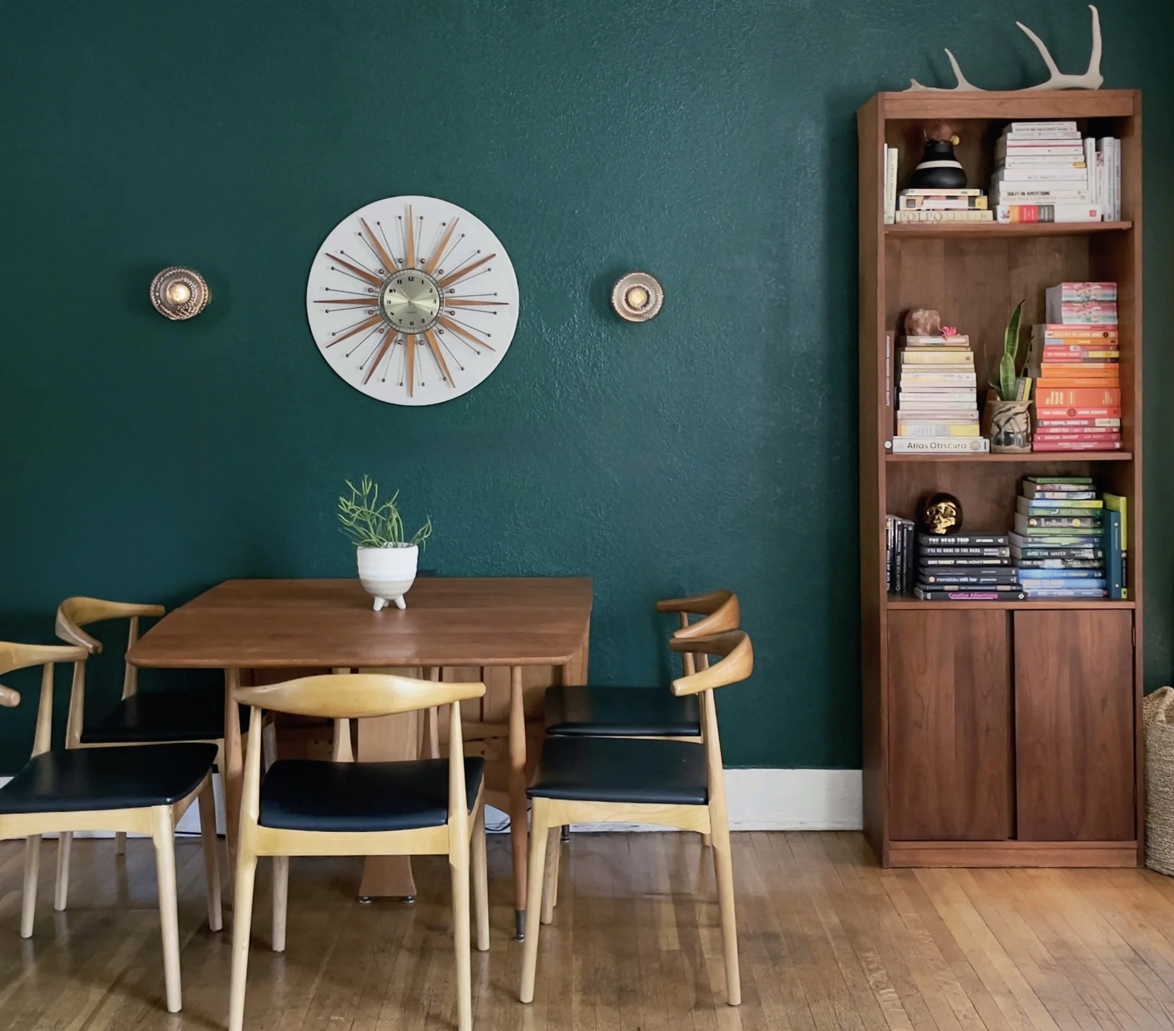 Echo Park mid-century dining room with rich green accent wall, vintage starburst clock, walnut dining table, wishbone chairs with black seats, custom wood bookcase, layered personal book collection, and warm California modern styling.