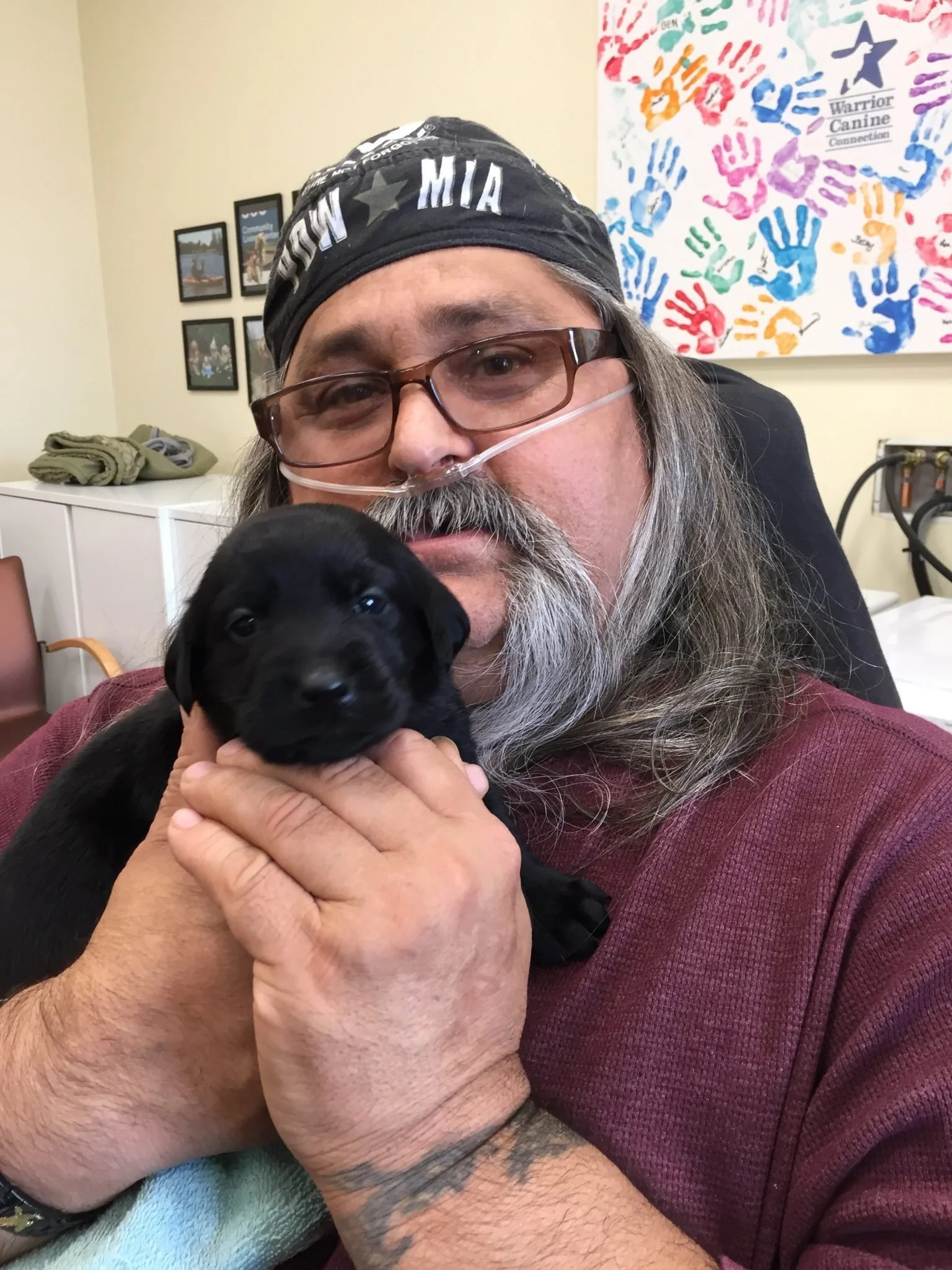 A man with glasses and a bandana holding a black puppy inside a room.